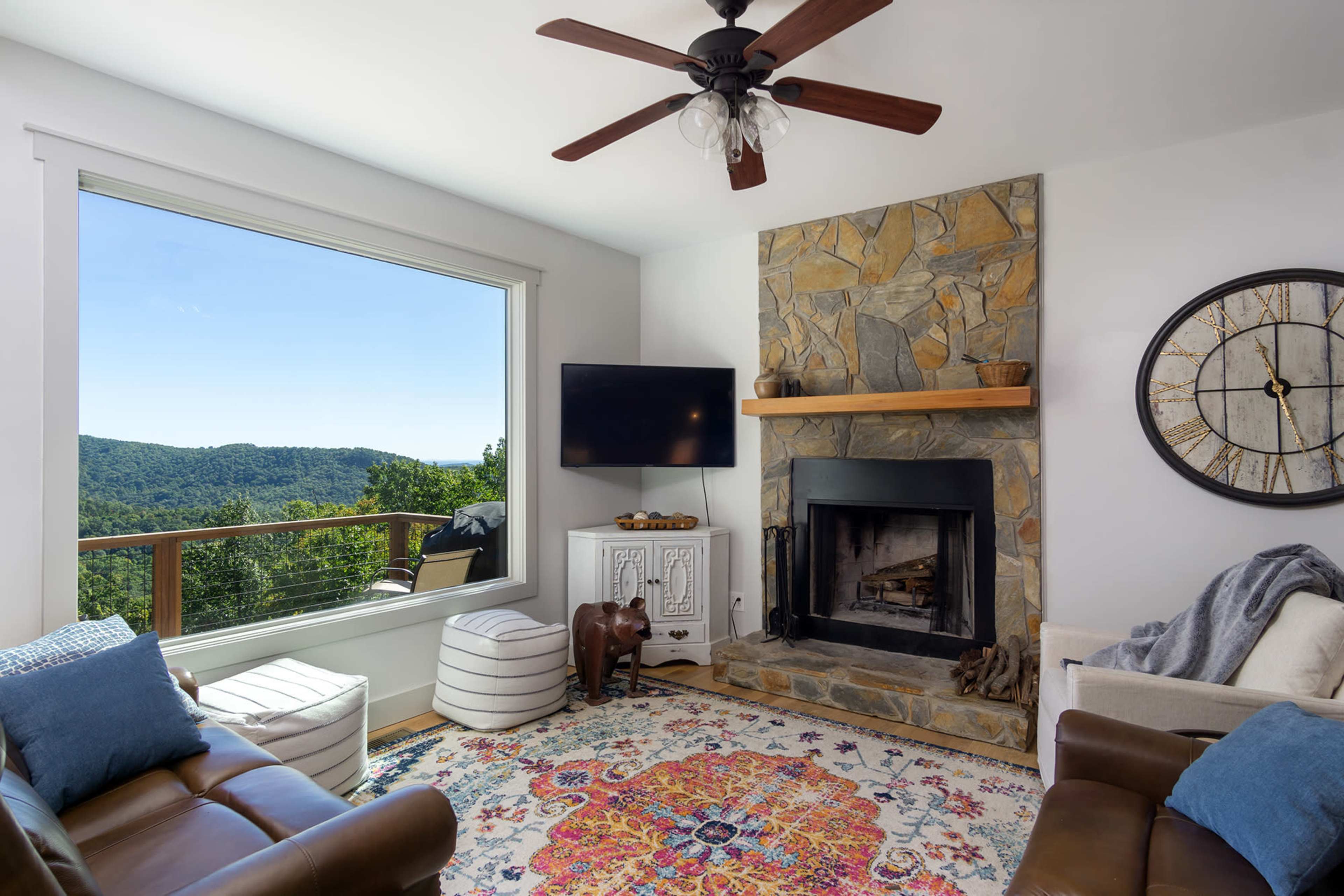 A living room features a stone fireplace, a television mounted on the wall, a large window with a mountain view, and decorative furnishings.