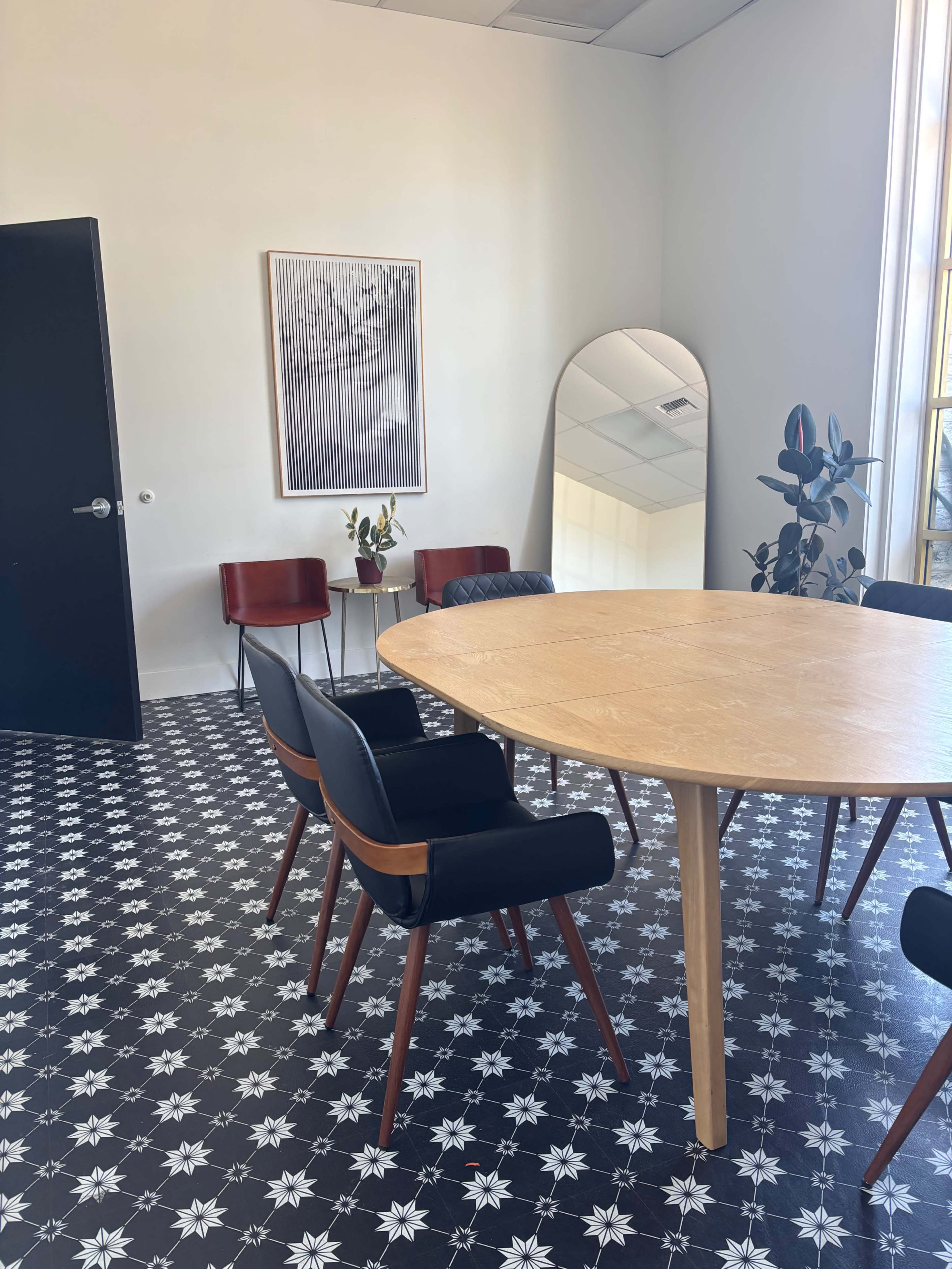 A modern meeting room features a round wooden table surrounded by black and wooden chairs, with a large mirror and patterned flooring.