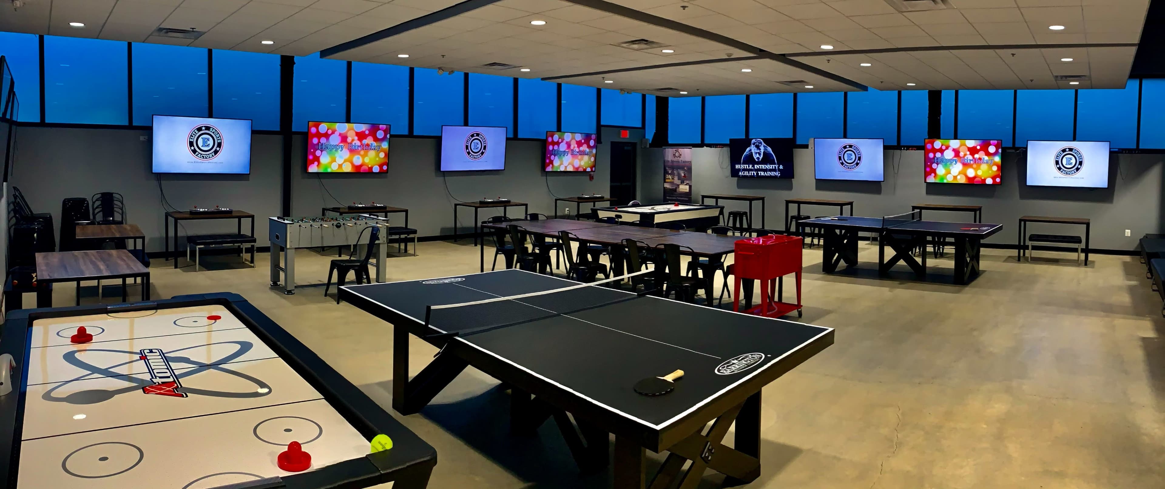 A modern game room featuring multiple air hockey tables, foosball tables, and large screens mounted on the walls, with large windows displaying a view of the outdoors.