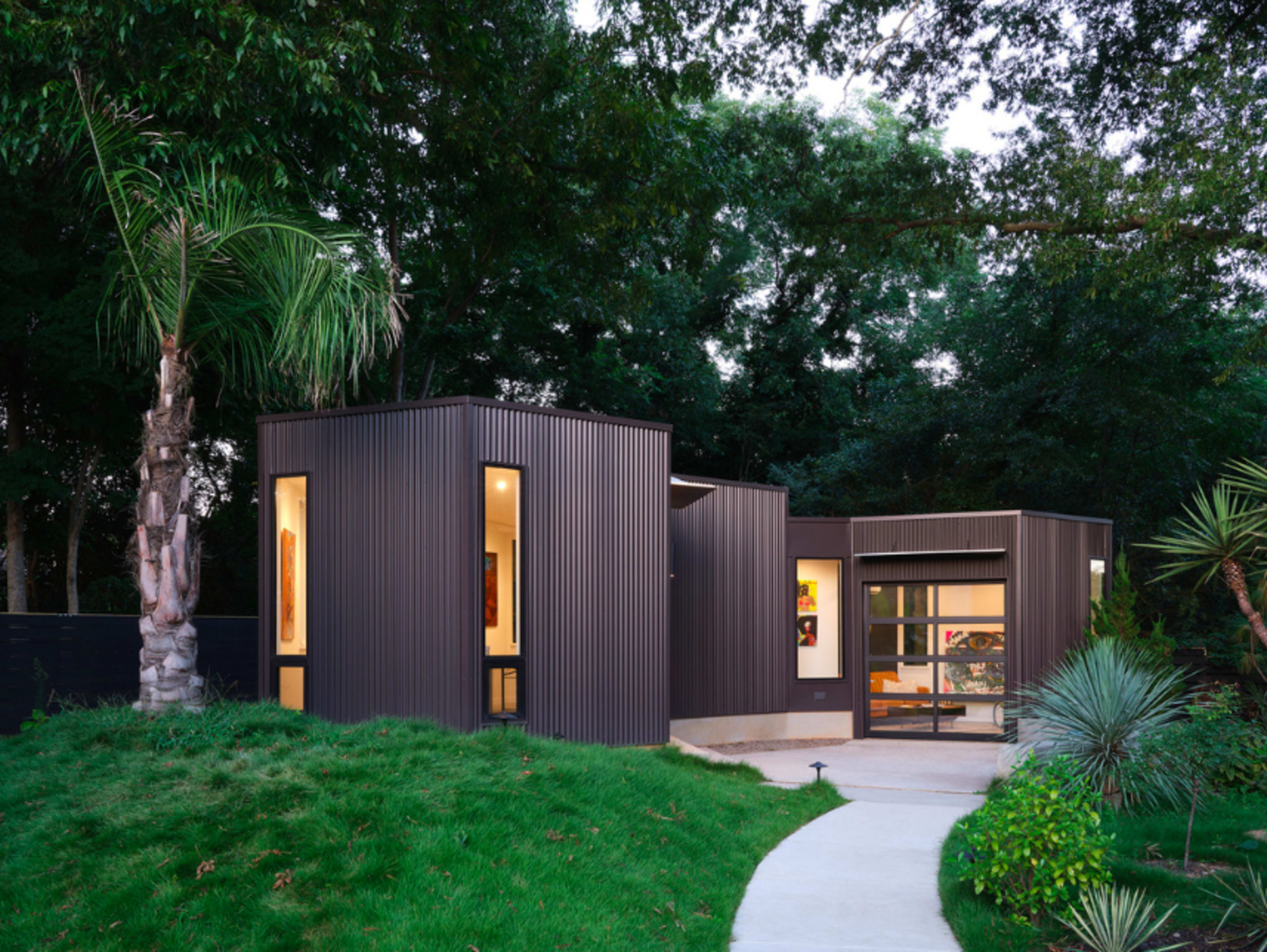A modern, angular house with dark metal siding is surrounded by lush greenery and features large windows.