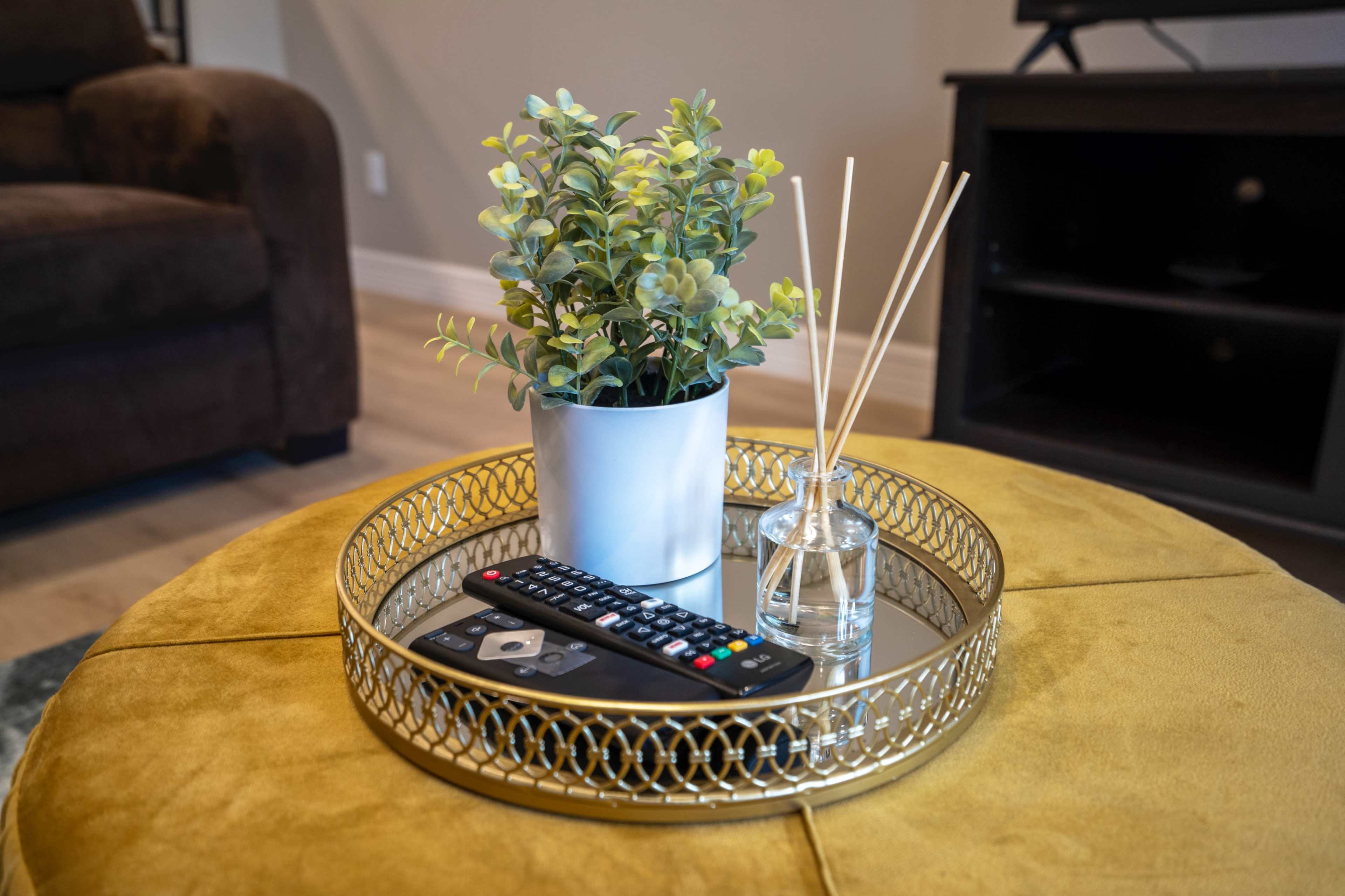 A round tray on a yellow ottoman holds a remote control, a small potted plant, and a glass diffuser with reeds.