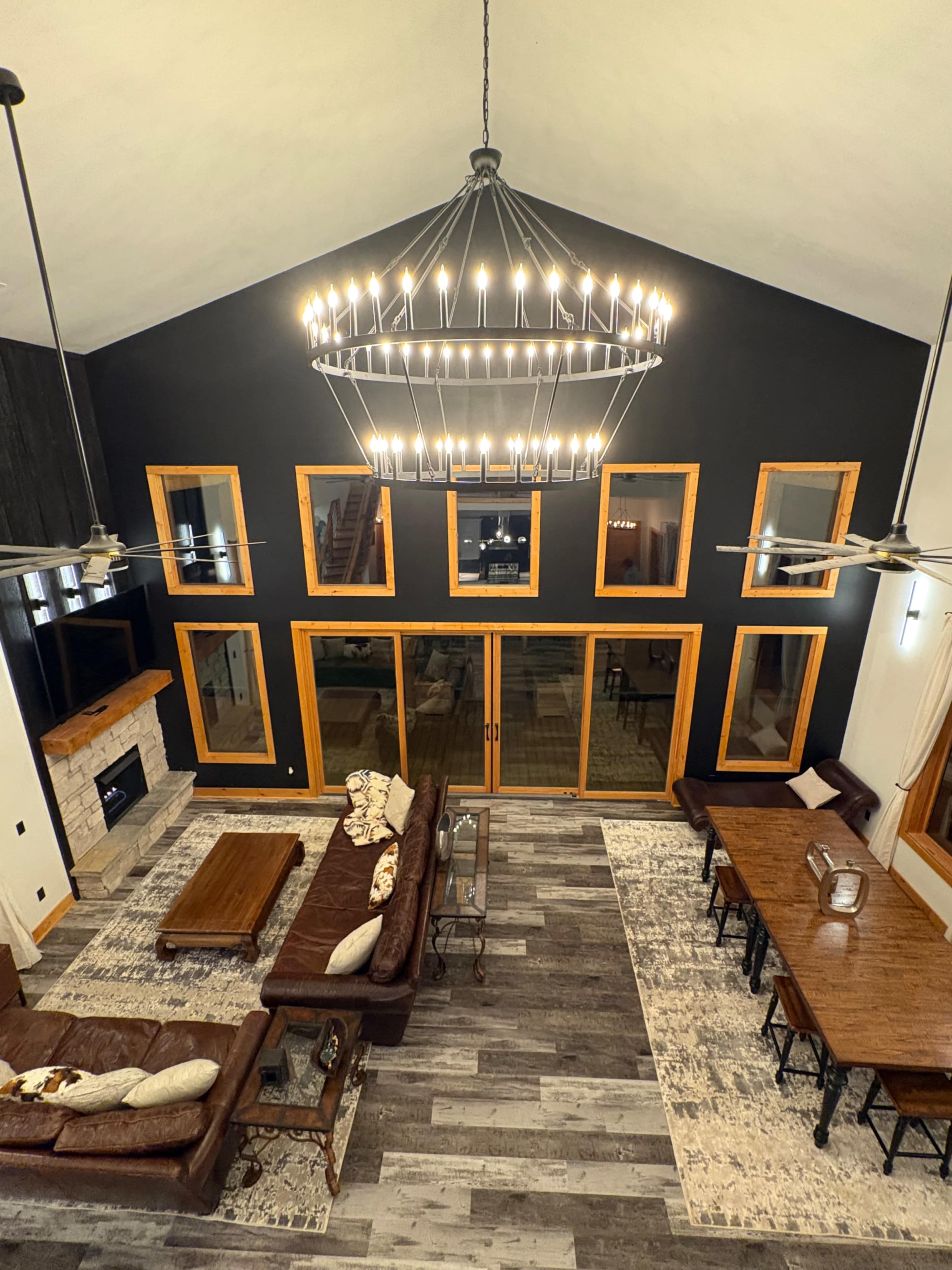 The image shows a large living area with a high ceiling, featuring a sectional sofa, a wooden dining table, large windows, and a prominent chandelier.