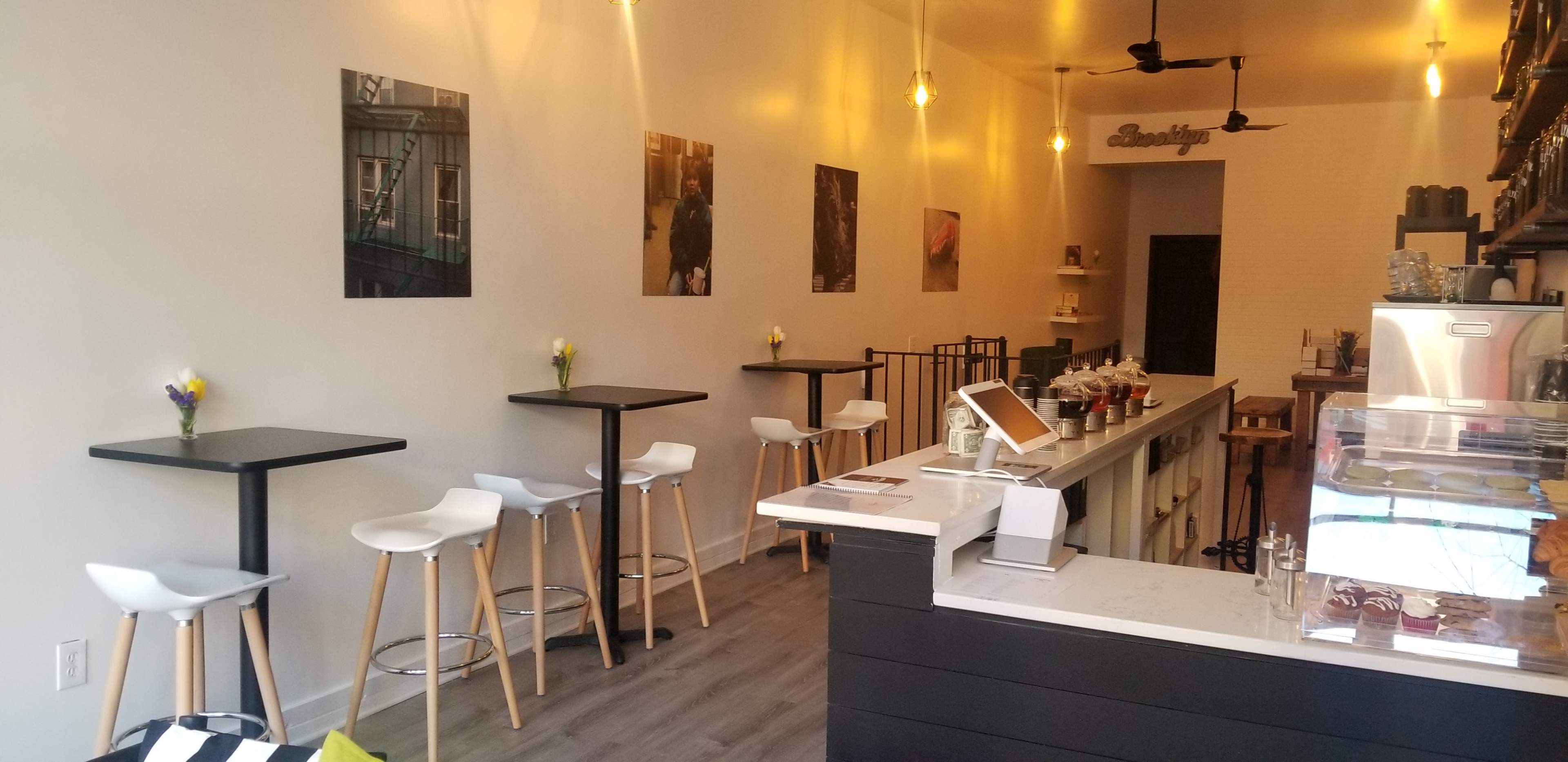 The image shows a modern café interior with a clean counter, stools, and wall art, illuminated by pendant lights.