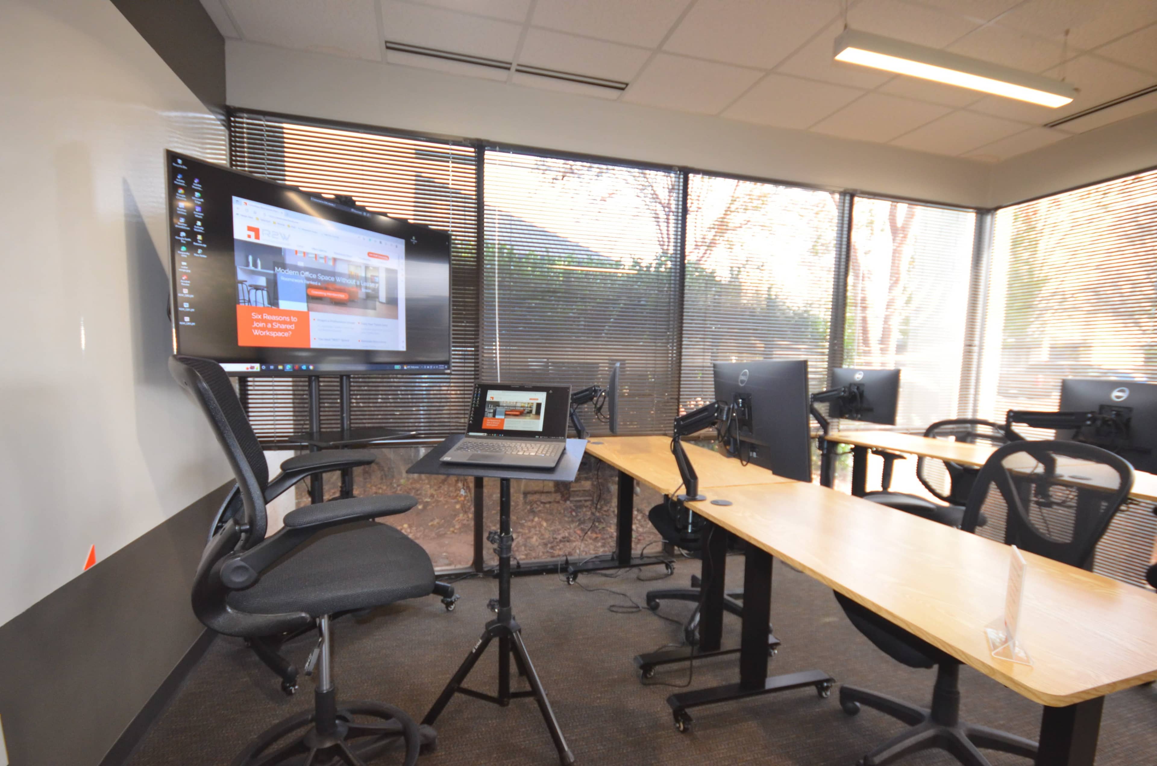 The image shows a modern office space featuring a computer monitor on a stand, several desks, and ergonomic chairs arranged near large windows.
