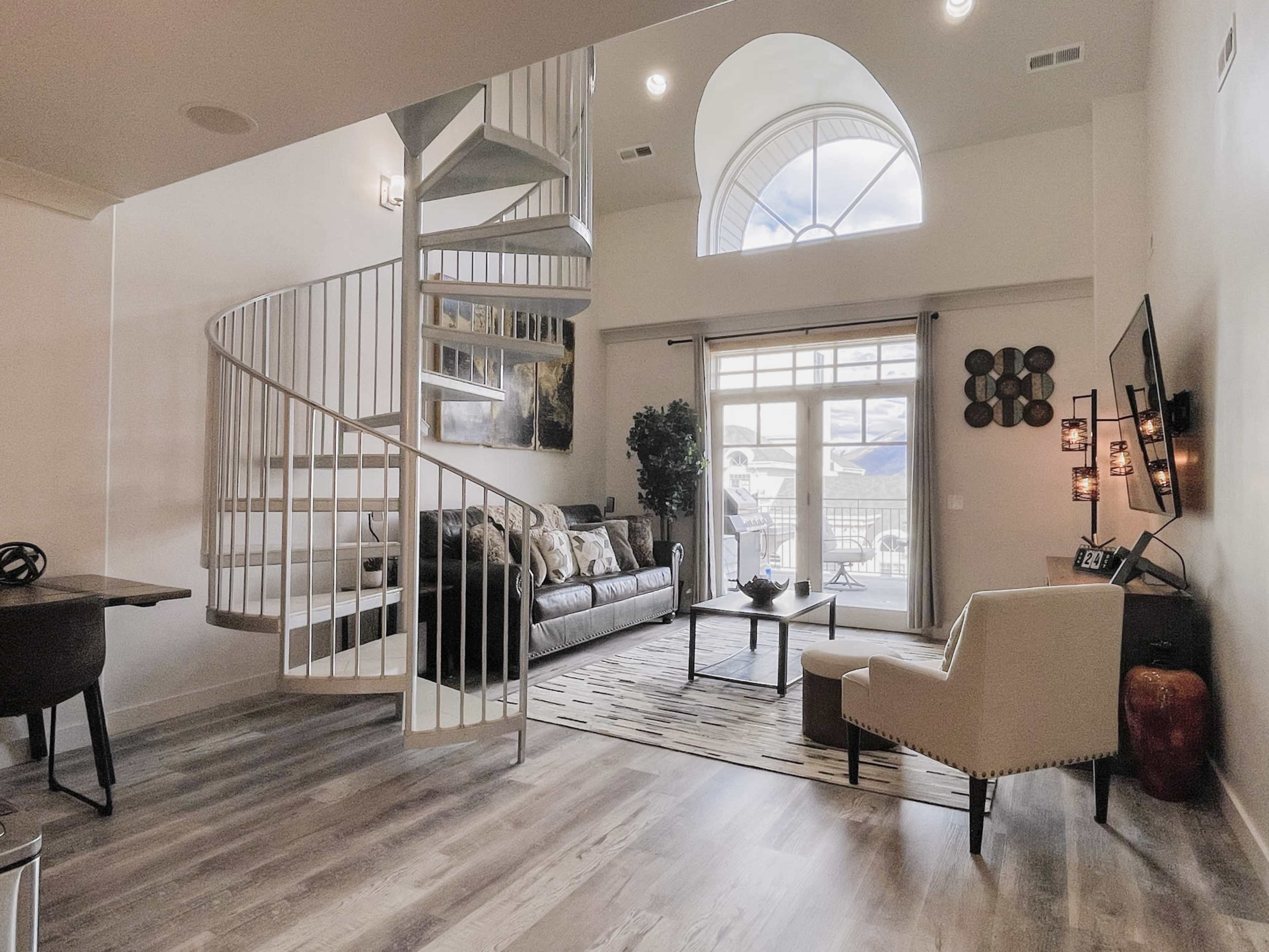 A modern living room with a spiral staircase, large windows, a comfortable seating area, and a light-colored rug.