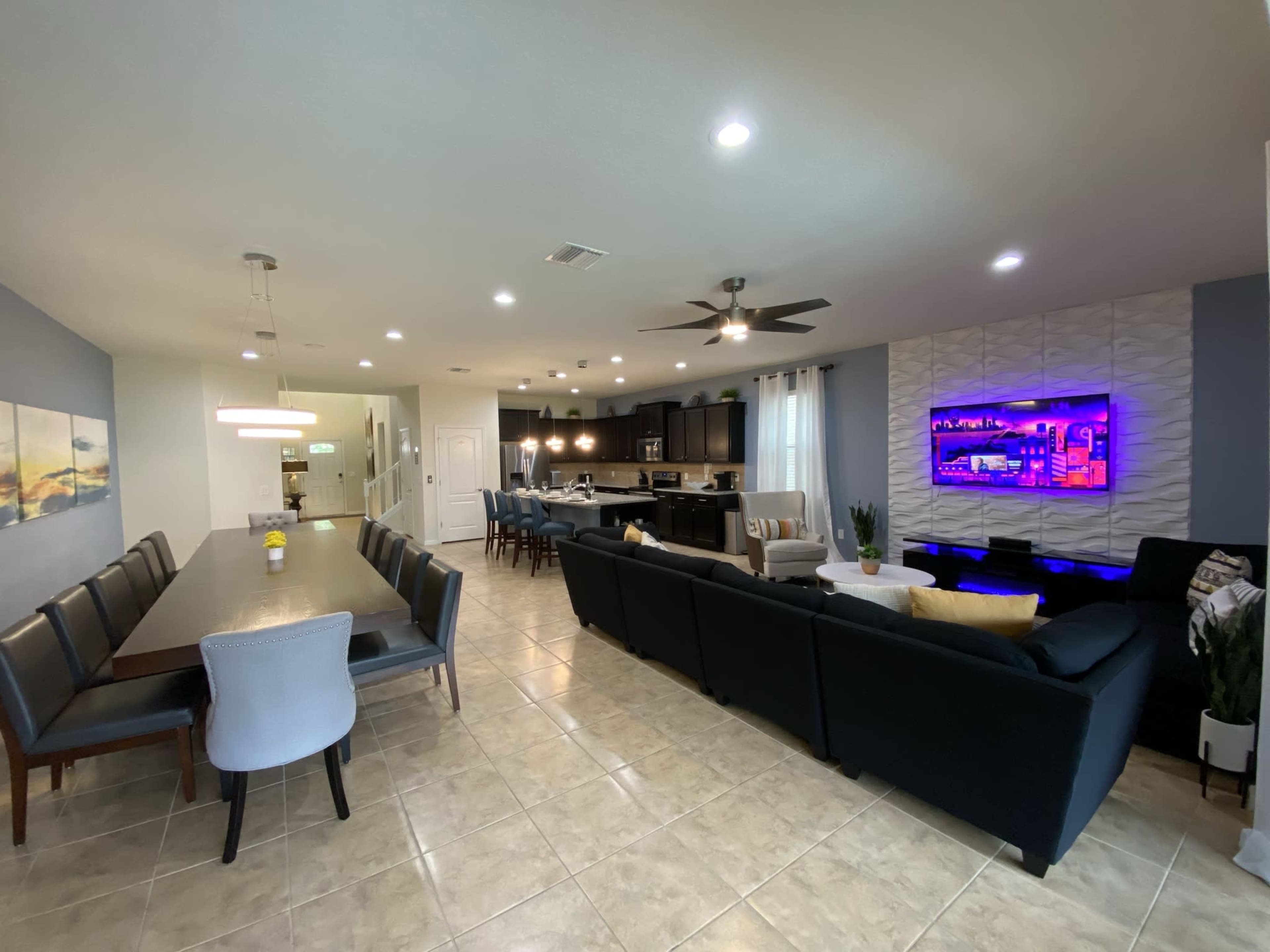 The image shows a modern open-concept living and dining area with a long dining table, a sectional sofa, and a kitchen visible in the background.