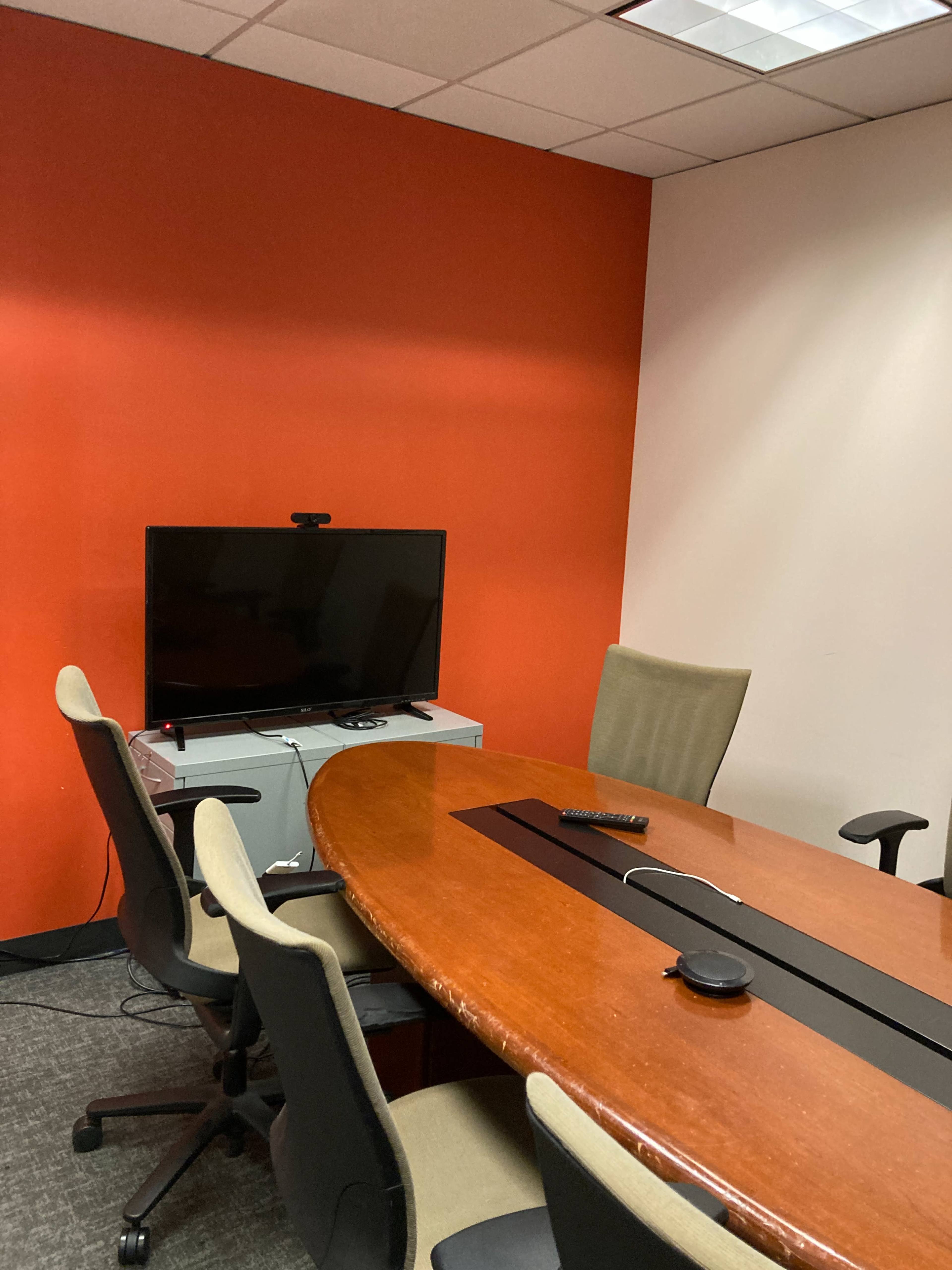 The image shows a small conference room with a round wooden table, several chairs, a television mounted on the wall, and an orange accent wall.