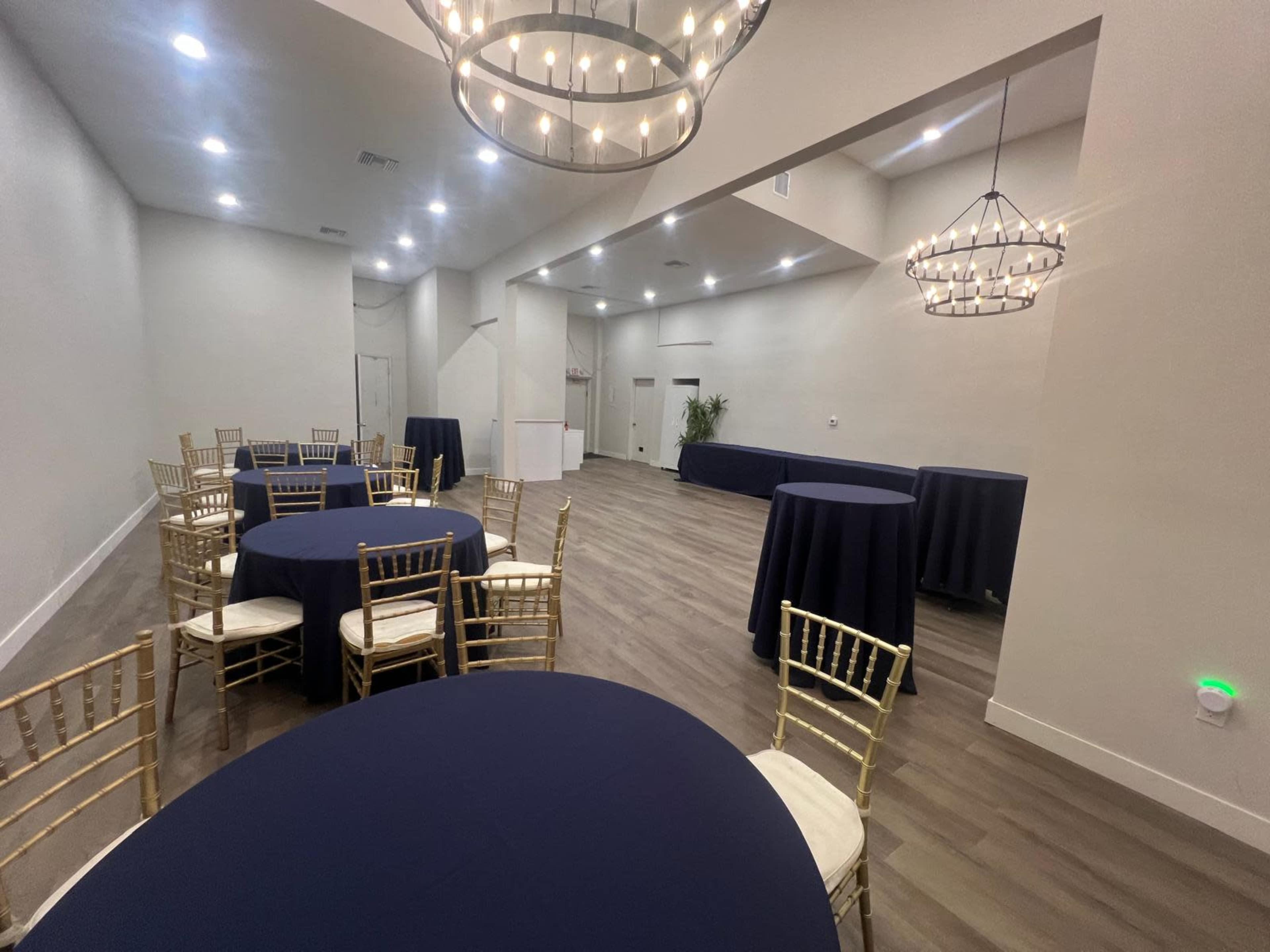 A spacious event room features several round tables covered with navy tablecloths and gold chairs, along with stylish chandeliers hanging from the ceiling.