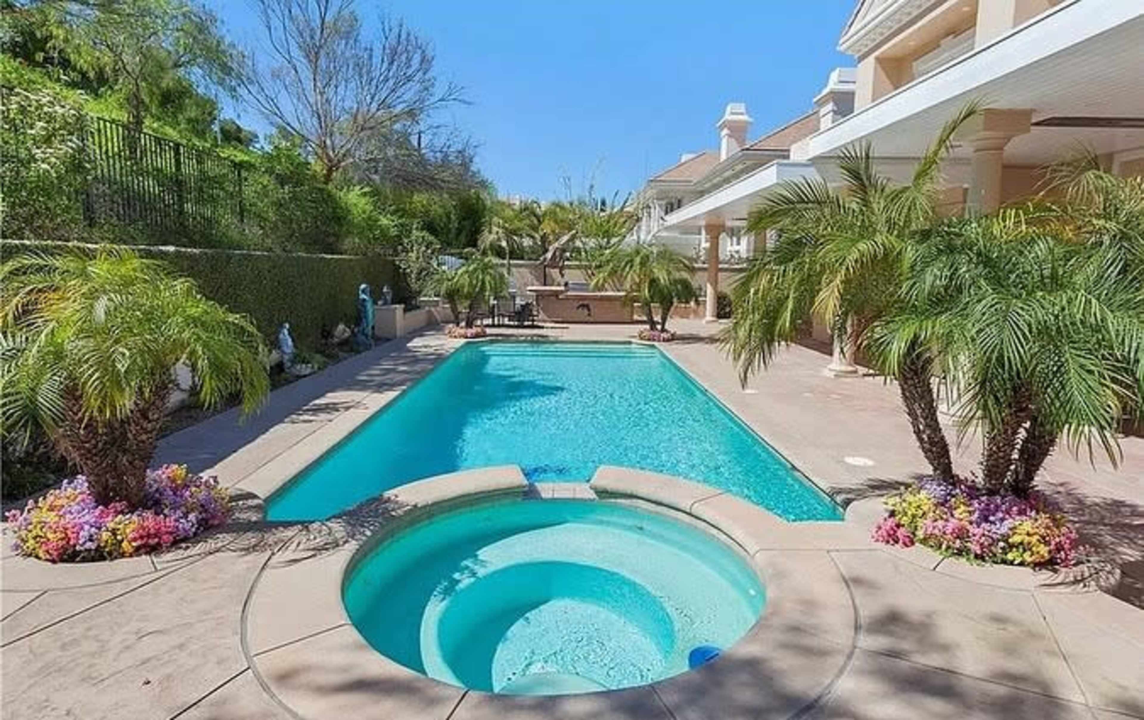 The image shows a backyard pool area featuring a large swimming pool with a connected hot tub, surrounded by palm trees and colorful flower beds.