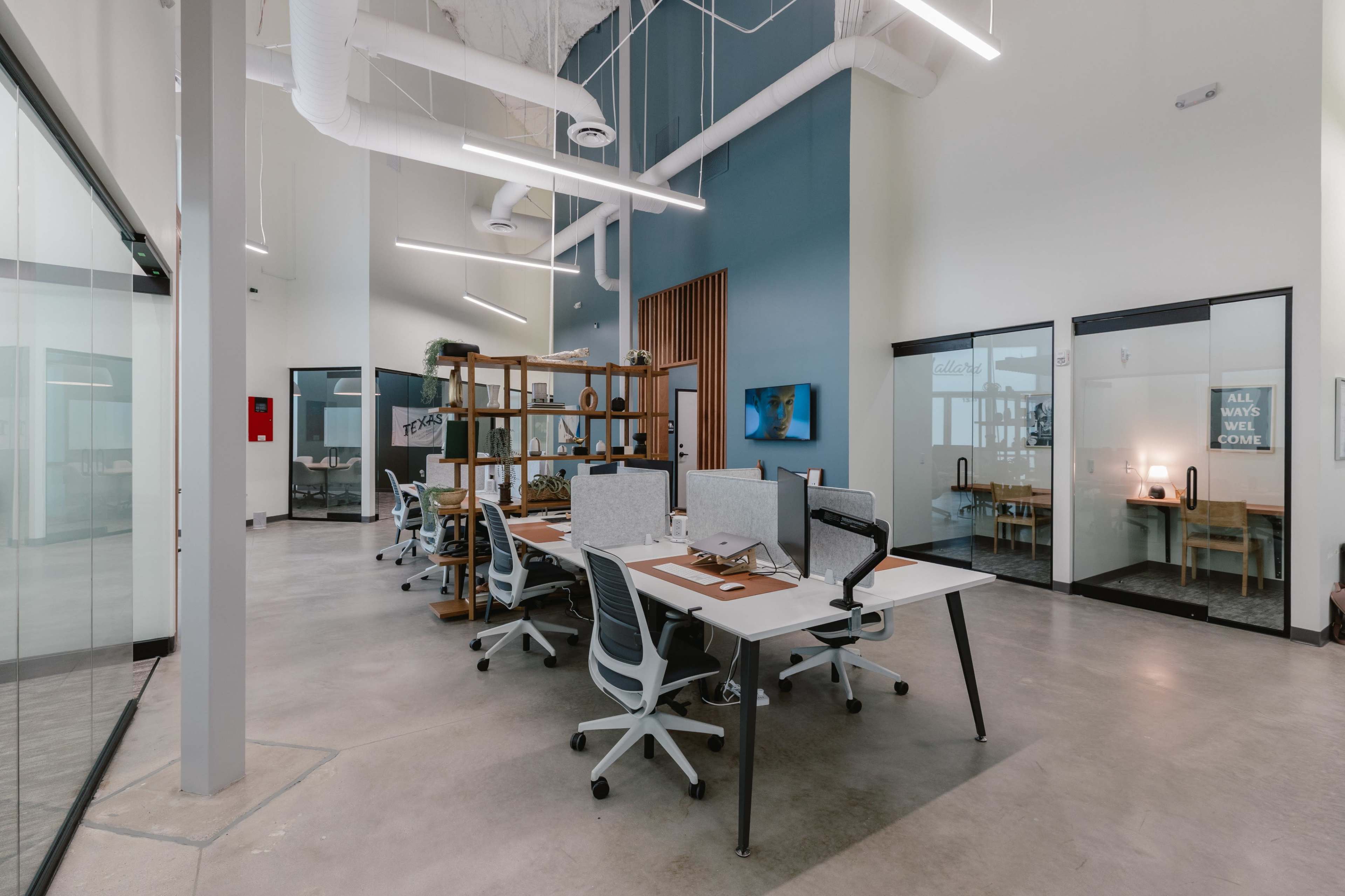 The image depicts a spacious office environment with modern desks and chairs arranged in a layout that encourages collaboration, featuring glass-walled meeting rooms and a neutral color scheme.