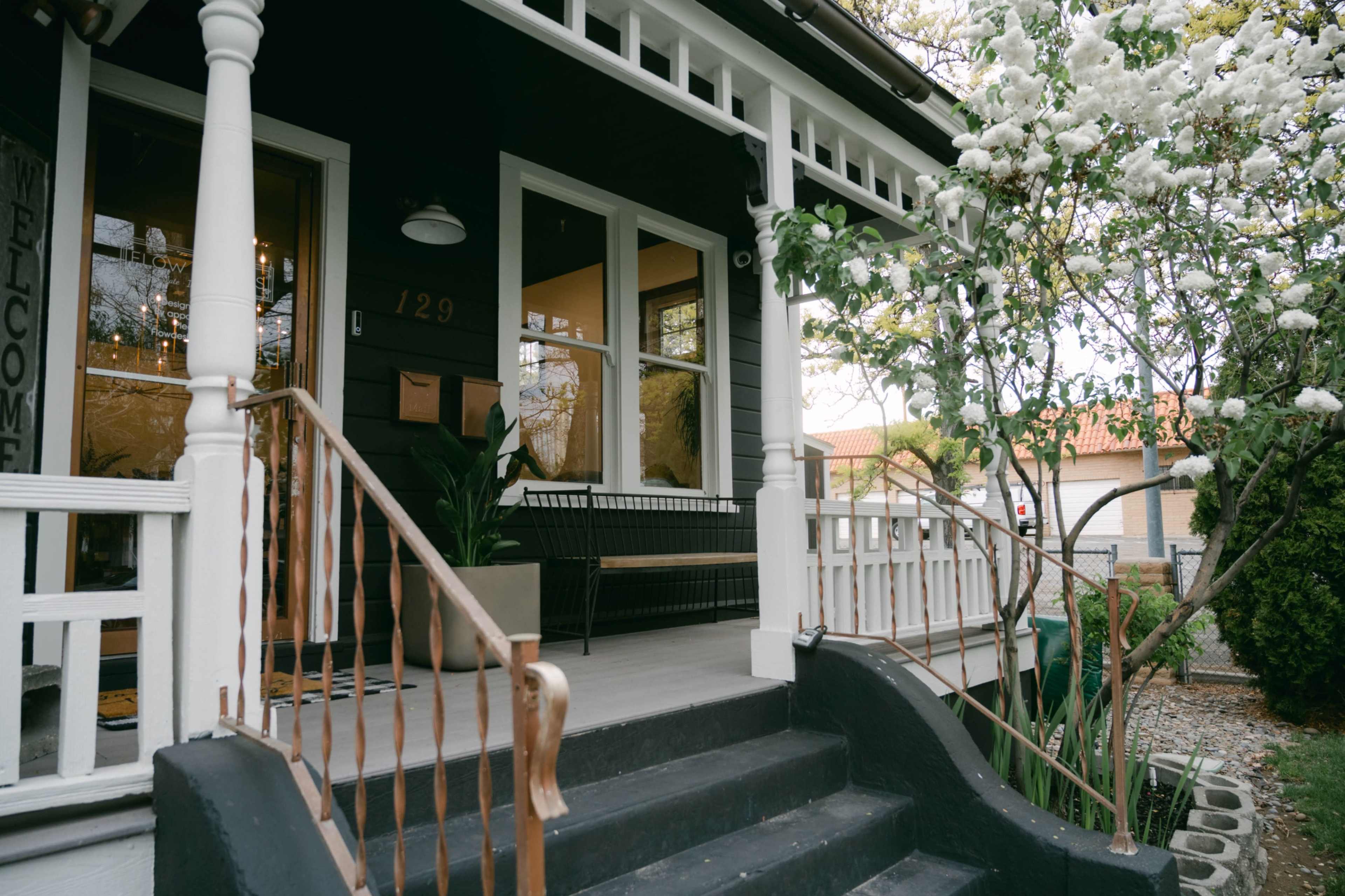 The image shows a black and white house with a front porch, featuring white railing and steps, surrounded by greenery and flowering trees.