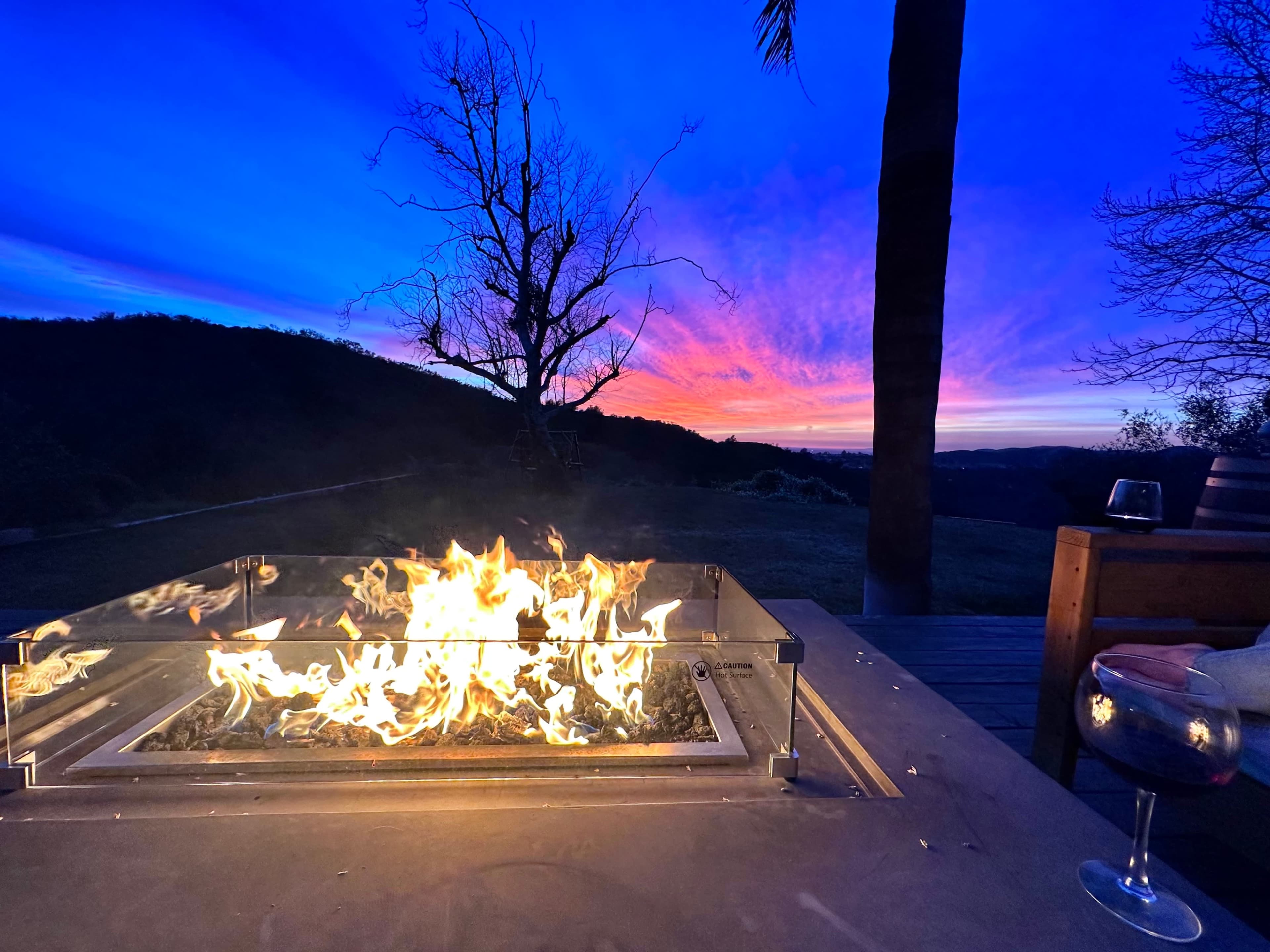 A fire pit with flames is lit in the foreground, while a colorful sunset with pink and blue skies is visible in the background.
