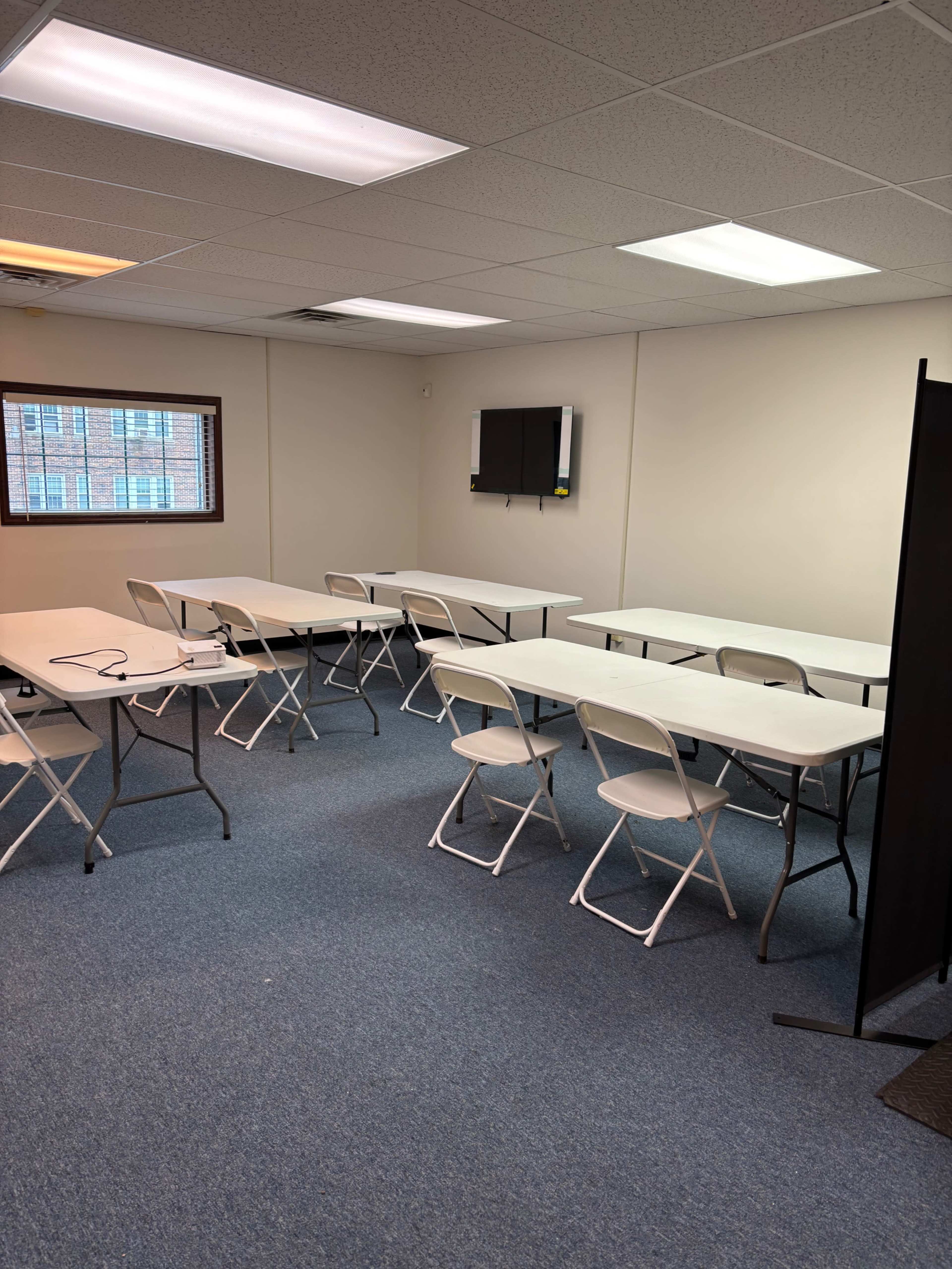 The image shows a rectangular room with several folding tables and chairs arranged neatly, a flat-screen TV mounted on the wall, and a partition on one side.