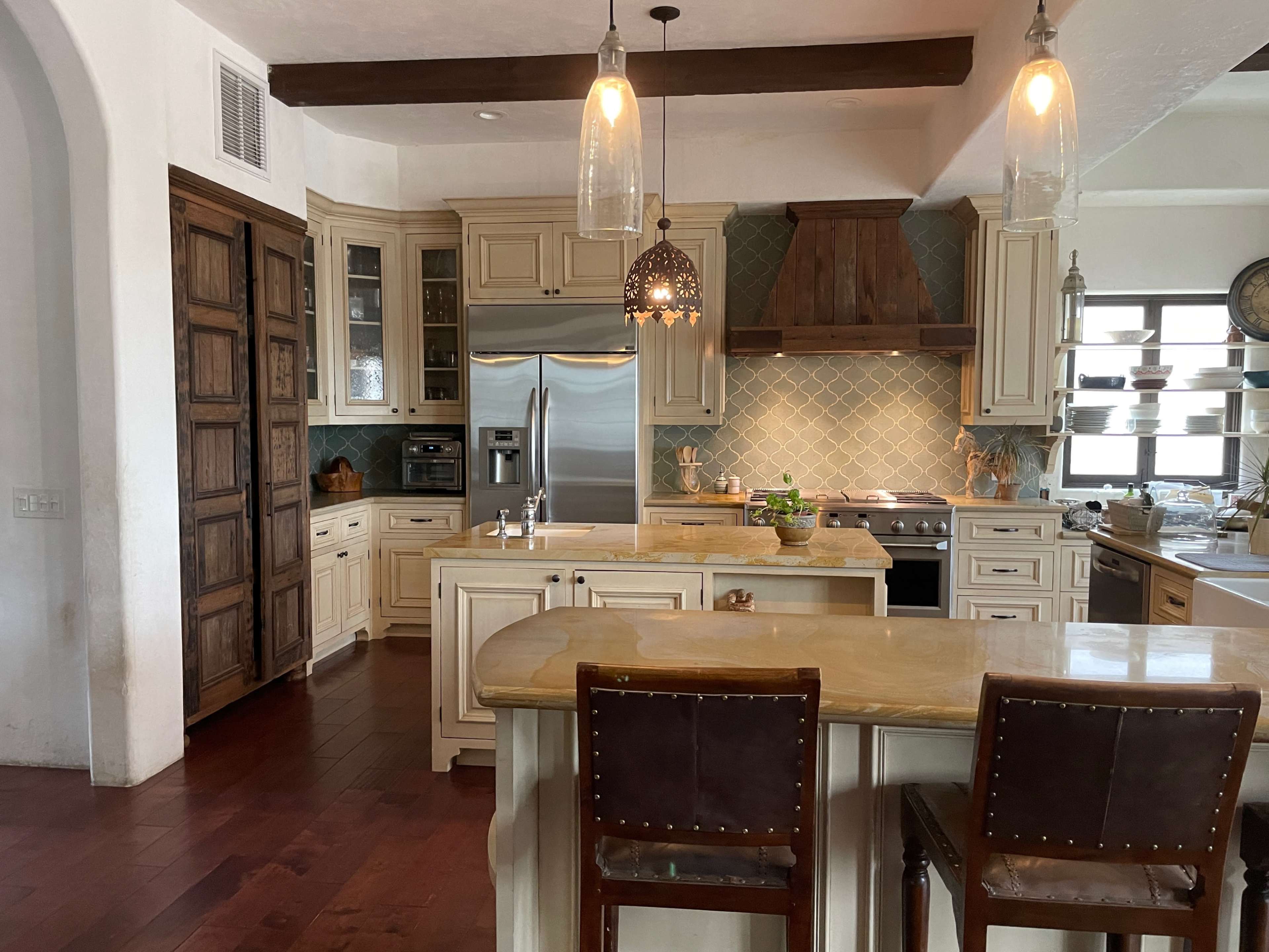 A kitchen with light-colored cabinetry, an island with two chairs, stainless steel appliances, and pendant lighting.
