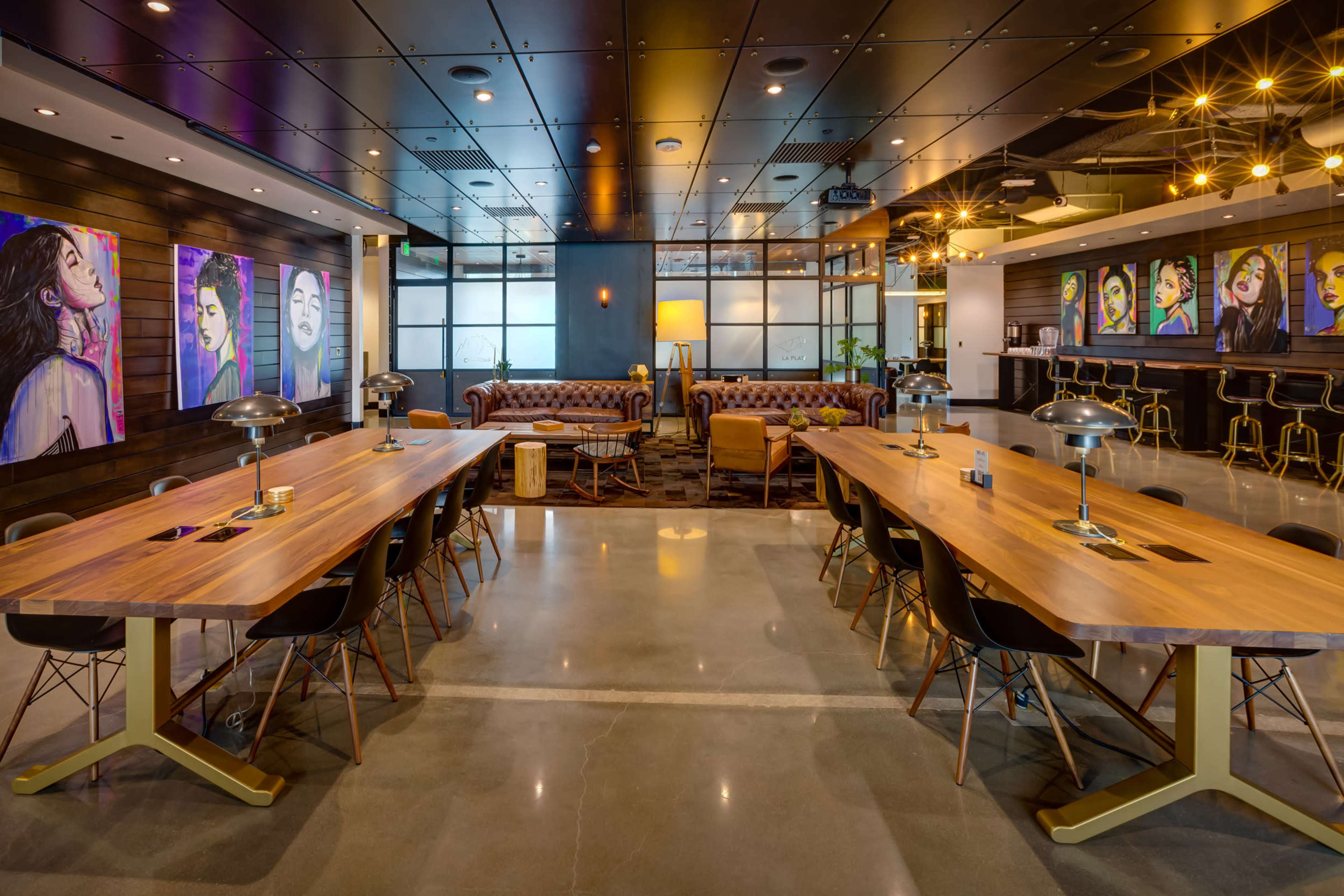 A modern coworking space features large wooden tables, leather sofas, and colorful artwork on the walls.