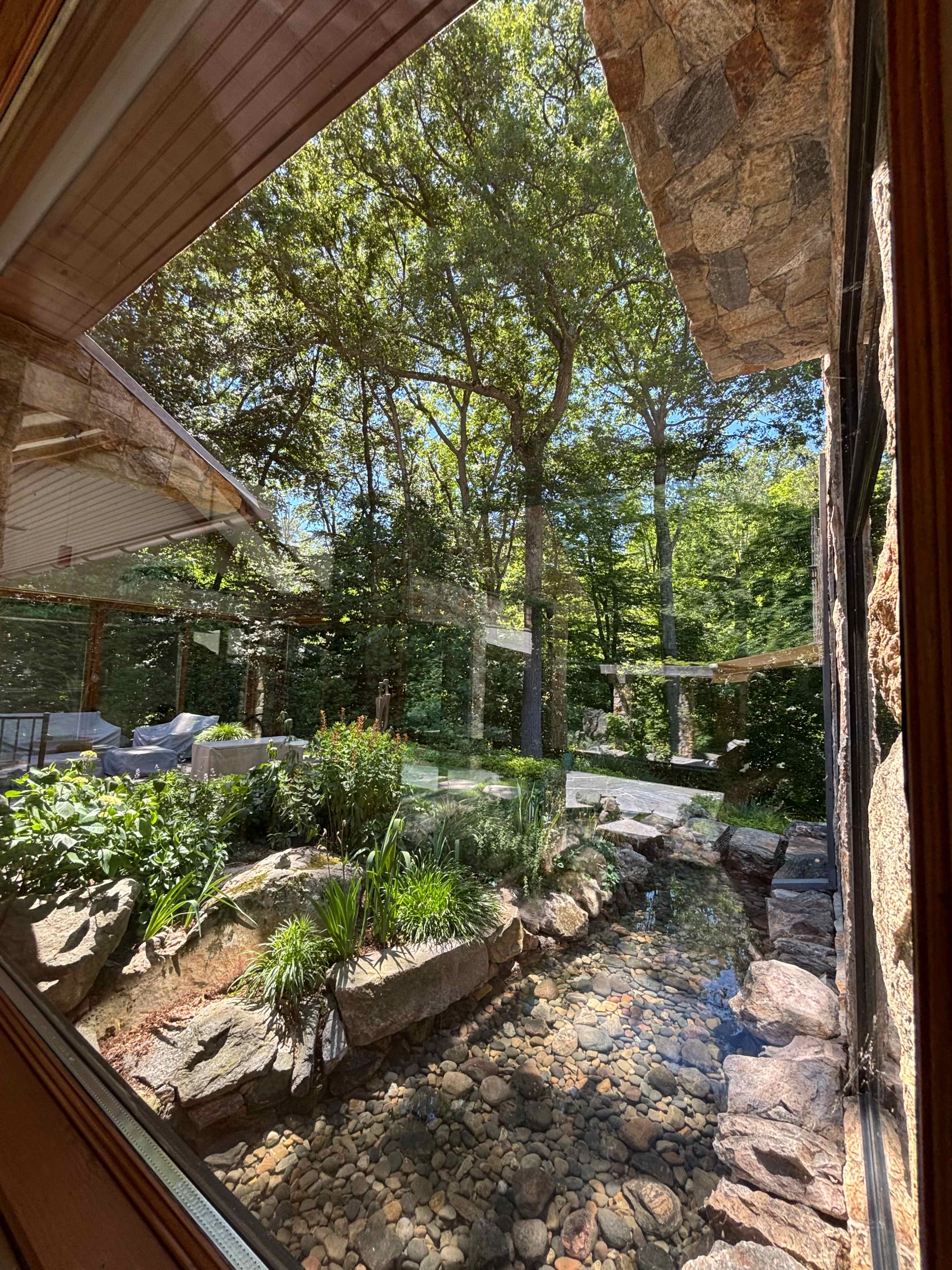 The image shows a view of a landscaped garden with a stone path, a small stream, and various plants, framed by a window of a stone-walled building.