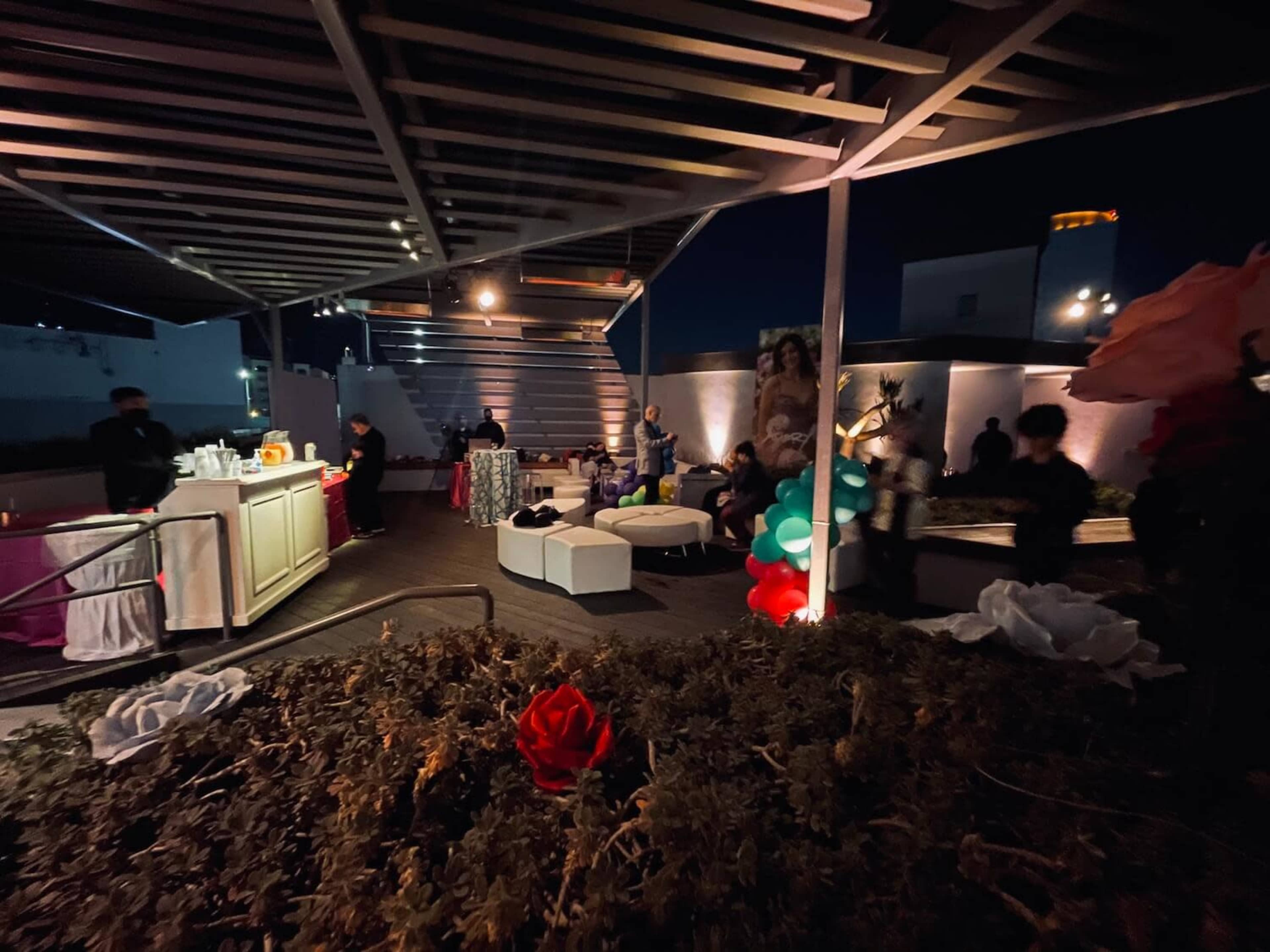 A dimly lit outdoor gathering features a bar area and guests socializing amidst decorative elements like balloons and flowers.