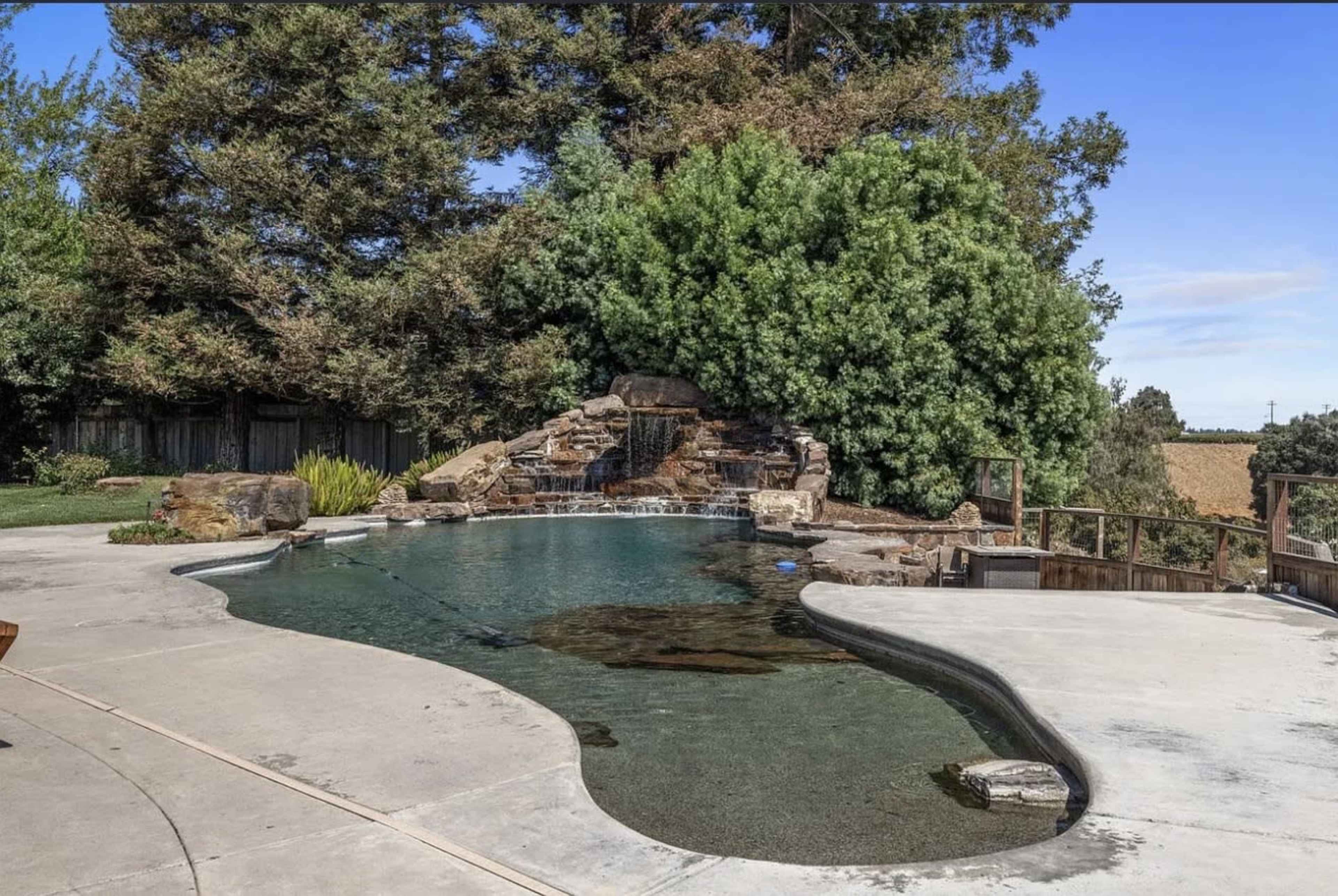 The image shows a swimming pool with a rock waterfall feature, surrounded by greenery and a landscaped area.