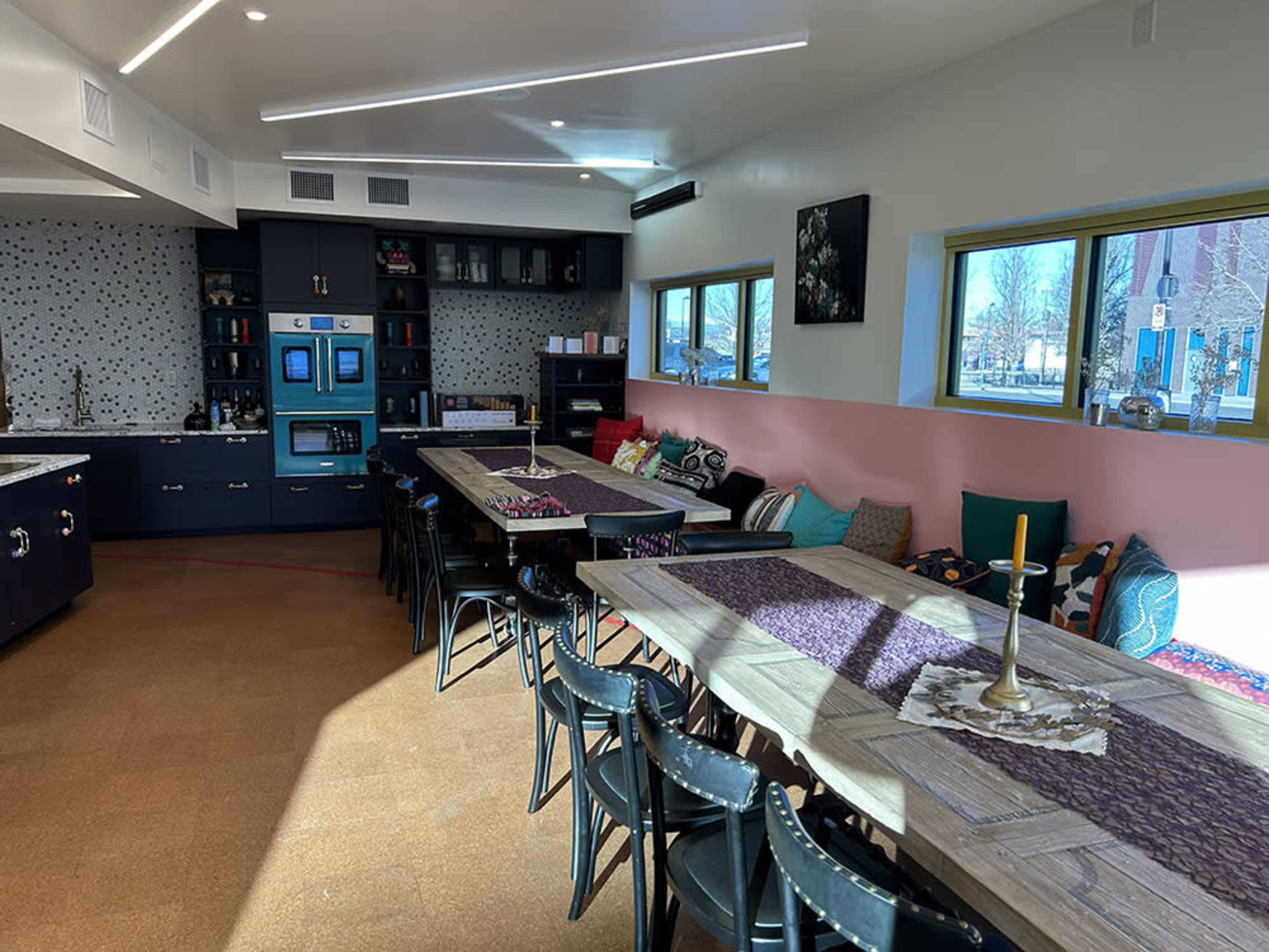 The image shows a modern kitchen and dining area with large wooden tables, colorful cushions, and well-organized cabinets.