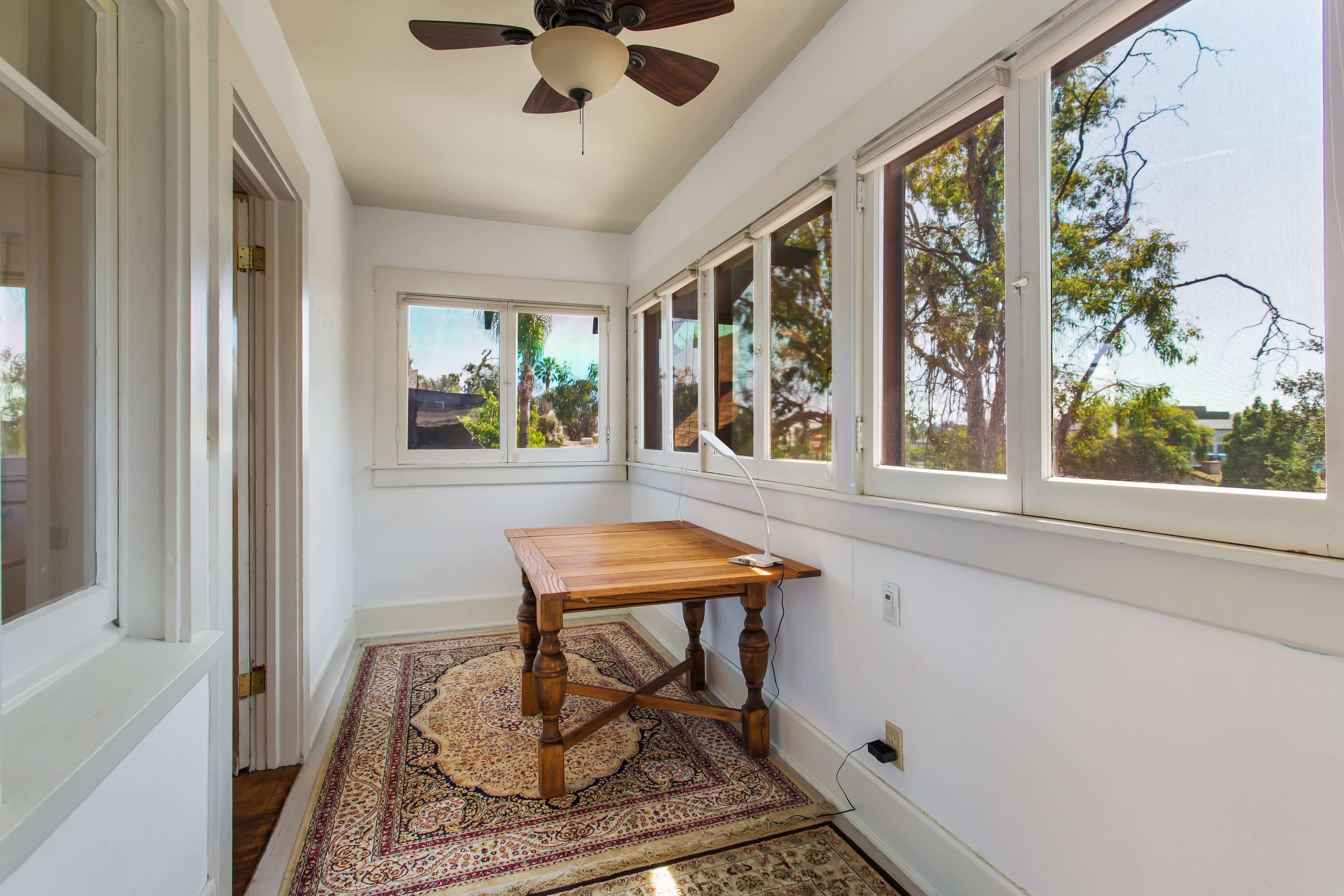 A sunlit room with large windows, a wooden table, and a decorative rug.