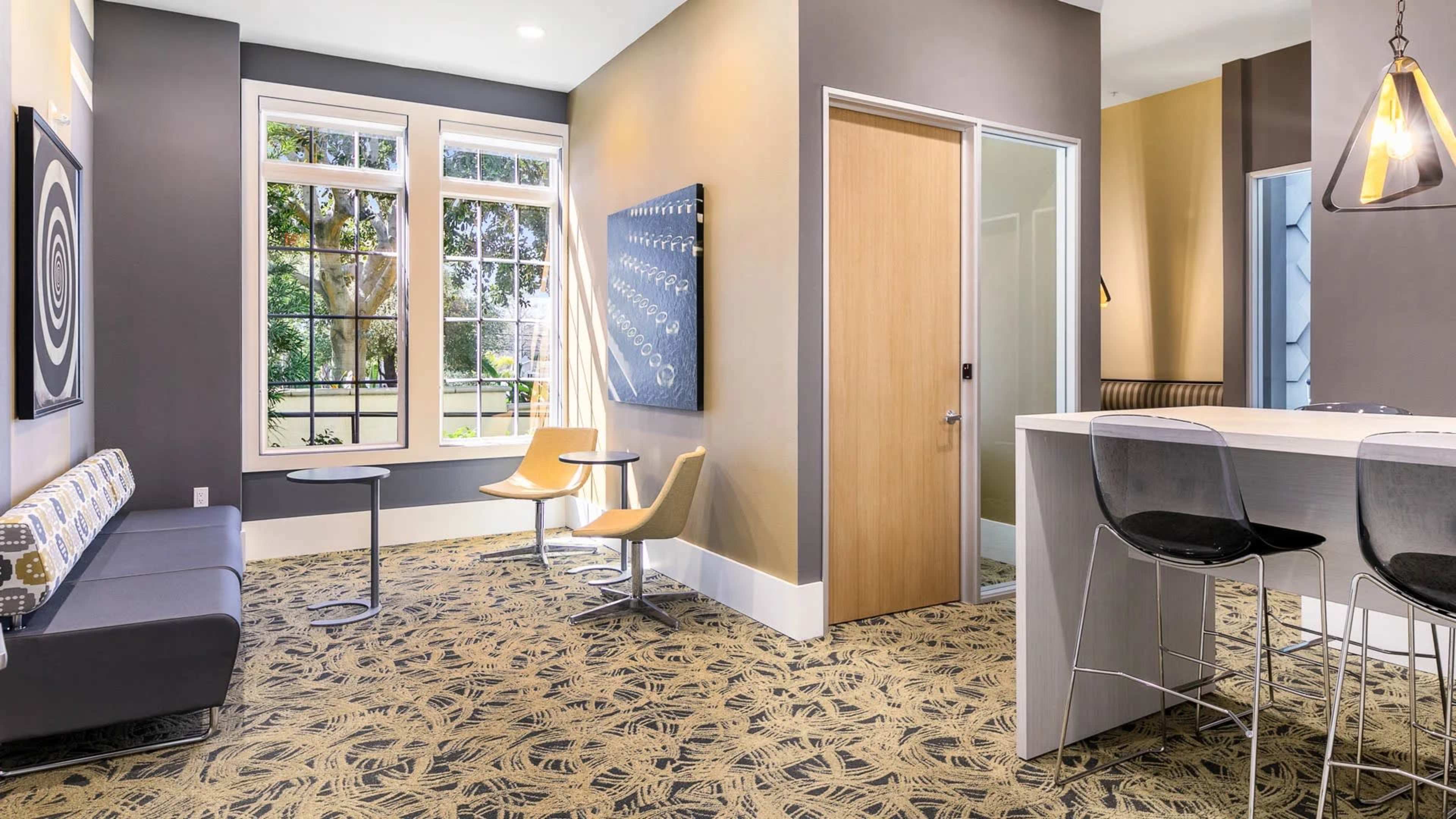 The image shows a modern lounge area with a gray couch, two round tables, and a kitchen bar in a well-lit room featuring large windows and a patterned carpet.