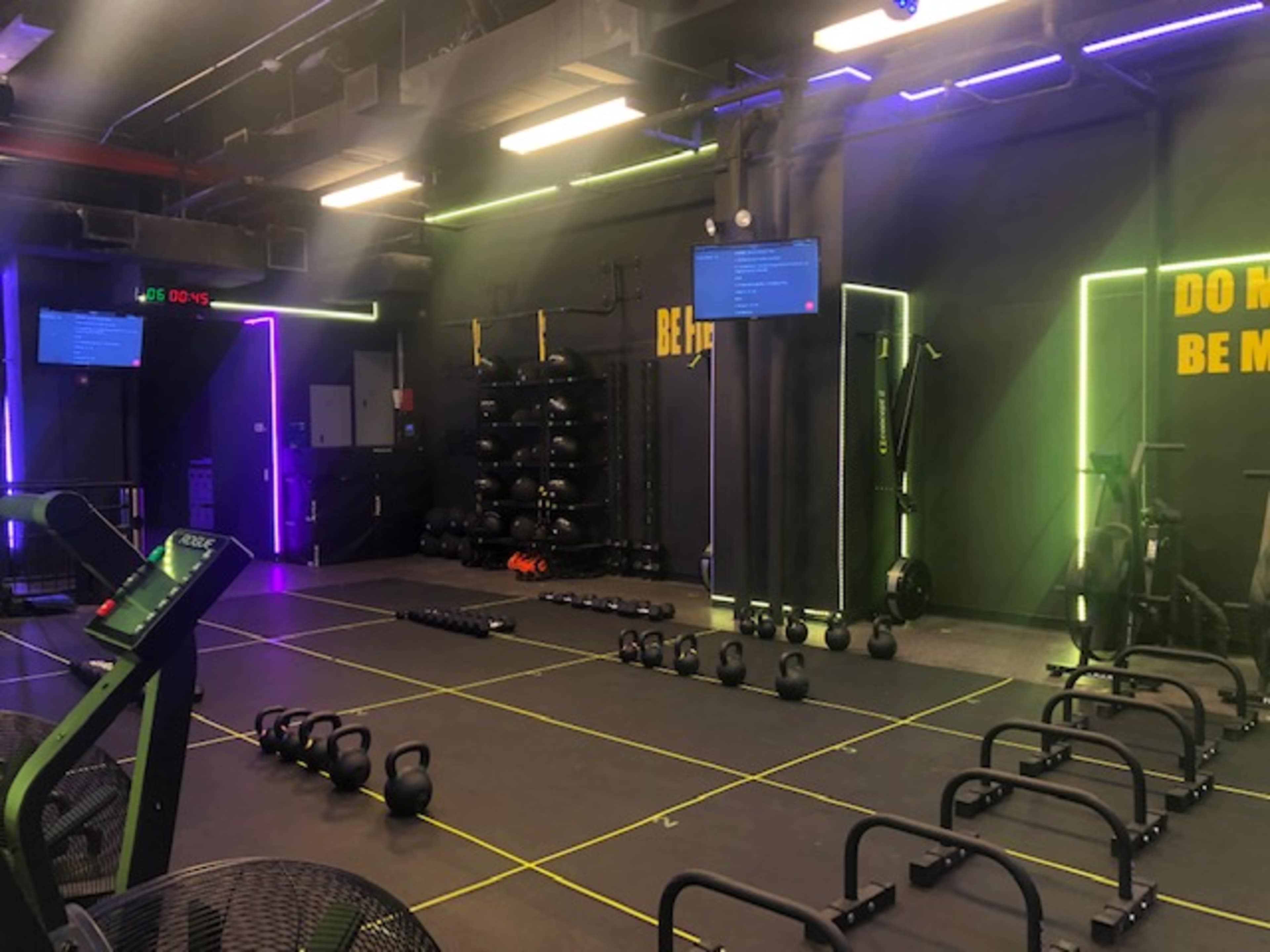 The image shows a modern gym interior equipped with kettlebells, exercise bikes, and a training area marked with grid lines under neon lights.