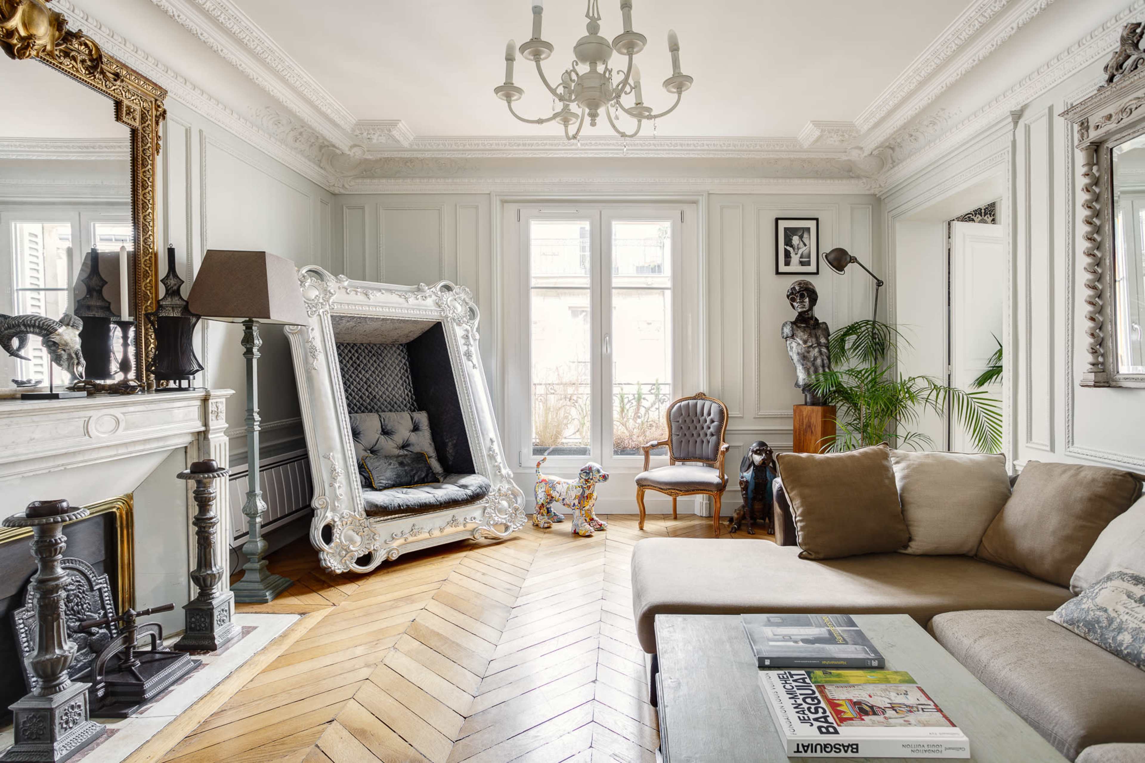 The room features a grand, ornate couch and a modern coffee table on a herringbone wood floor, complemented by large windows and decorative moldings.