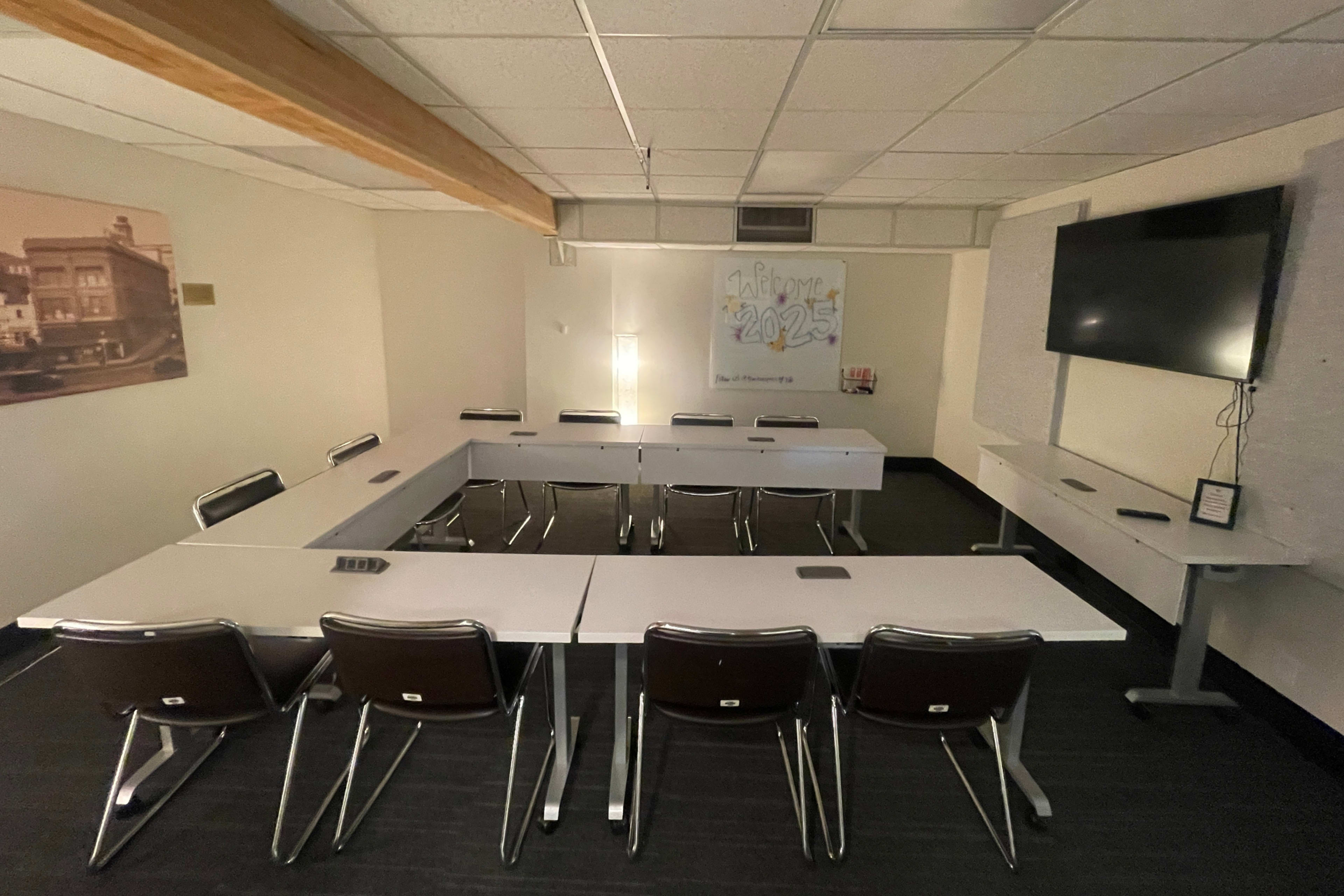 The image shows a meeting room set up with tables arranged in a U-shape, surrounded by chairs, and features a wall-mounted television and a whiteboard.