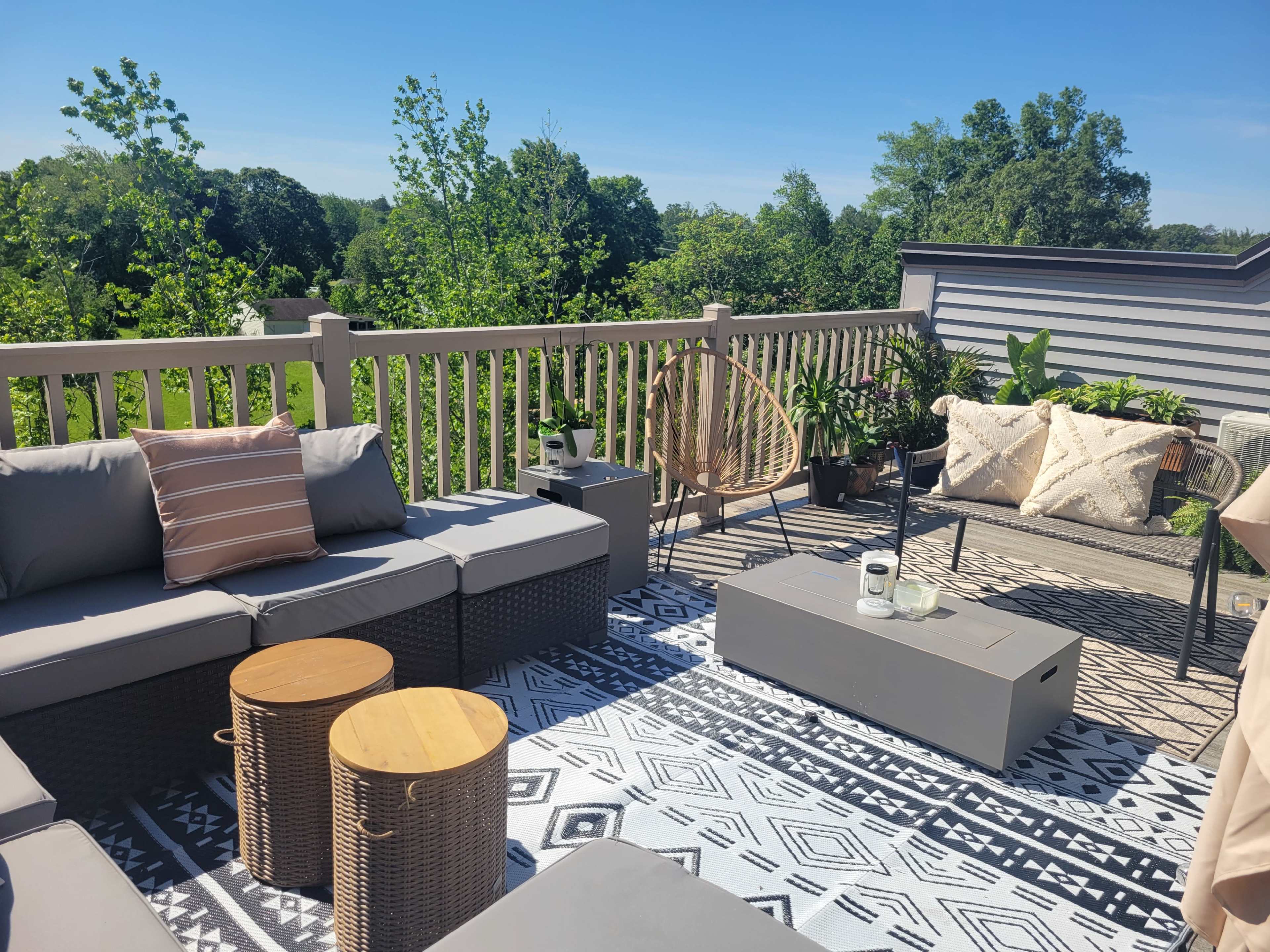 The image depicts a rooftop patio area featuring modern furniture, including a sectional sofa, wooden tables, and decorative plants, surrounded by greenery under a clear blue sky.