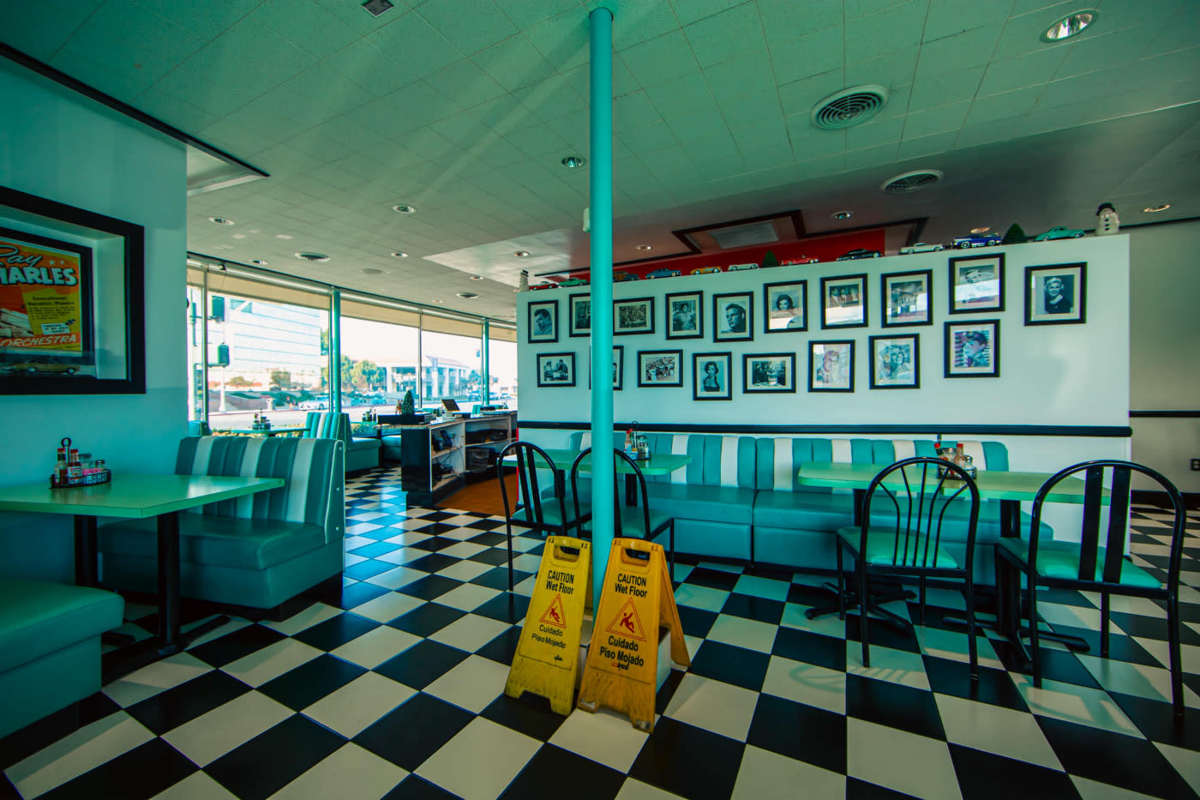 The image shows a retro diner interior with black and white checkered flooring, teal booths, and a row of framed photographs on the wall.