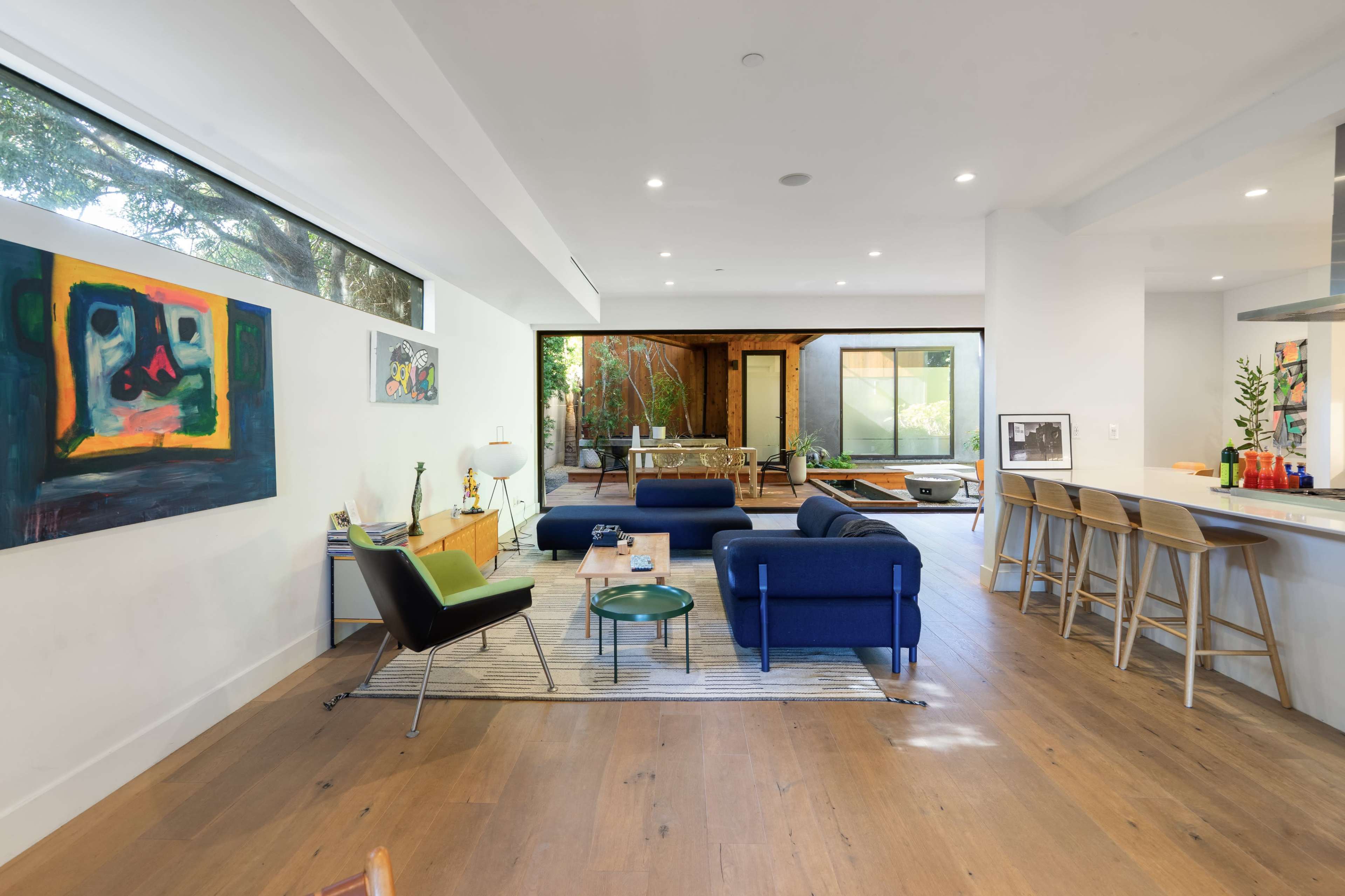 The image shows a modern living room with large windows, colorful artwork, and a view of an outdoor space.