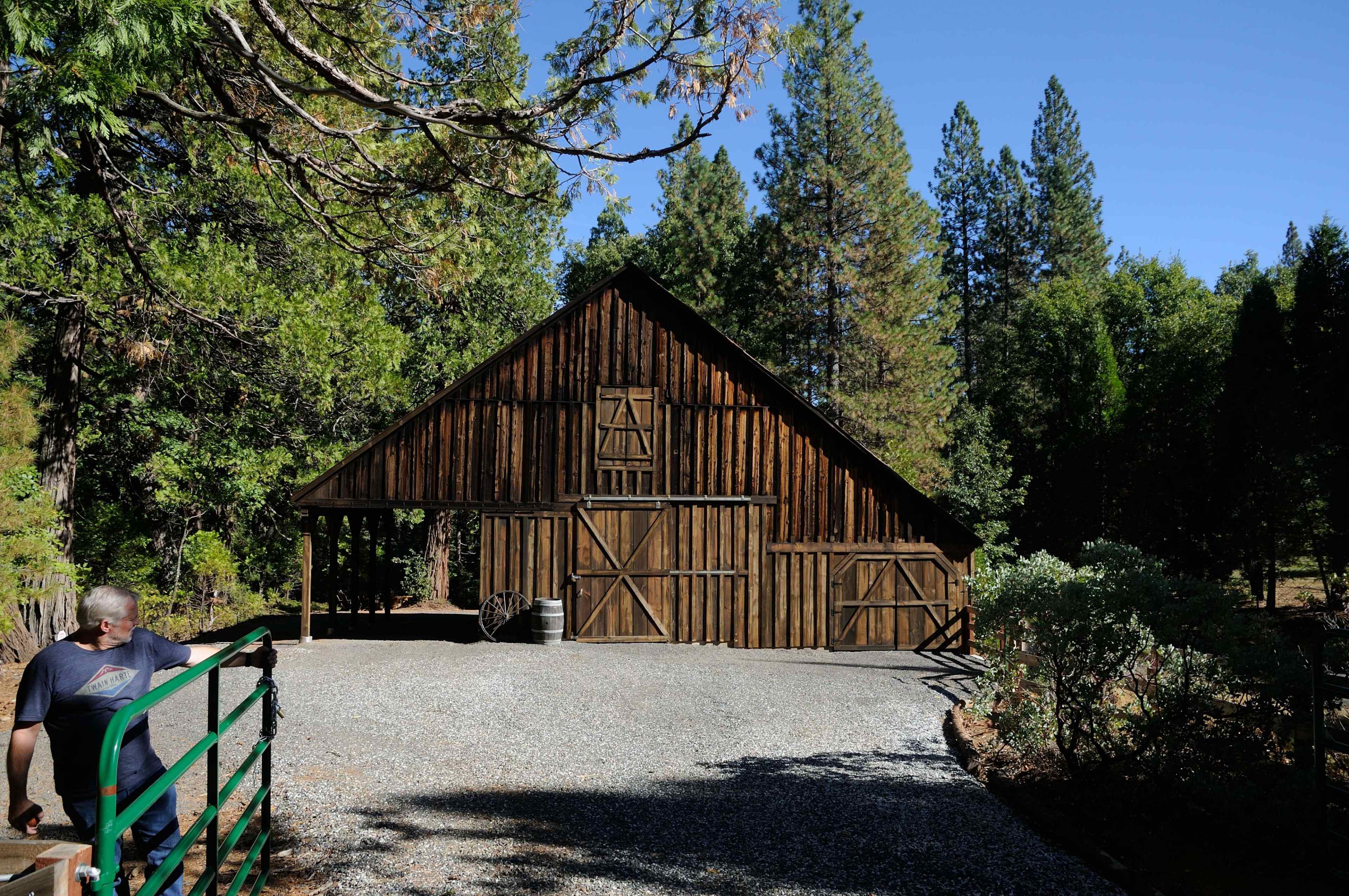 A wooden barn with a gabled roof is situated among tall trees and gravel pathways.