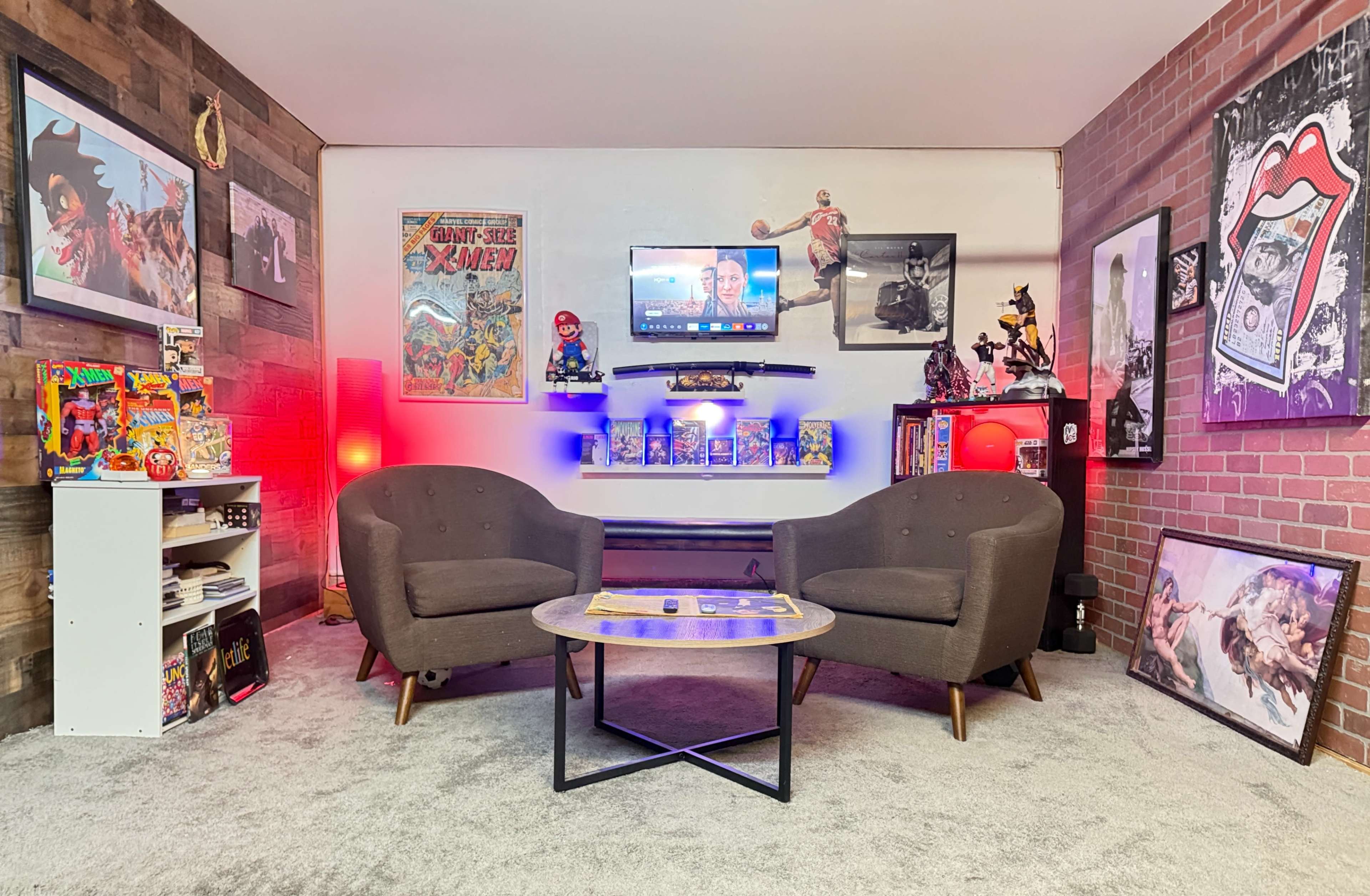 The image shows a cozy entertainment room featuring two gray upholstered chairs, a circular coffee table, wall art, collectibles, and a television mounted on the wall.