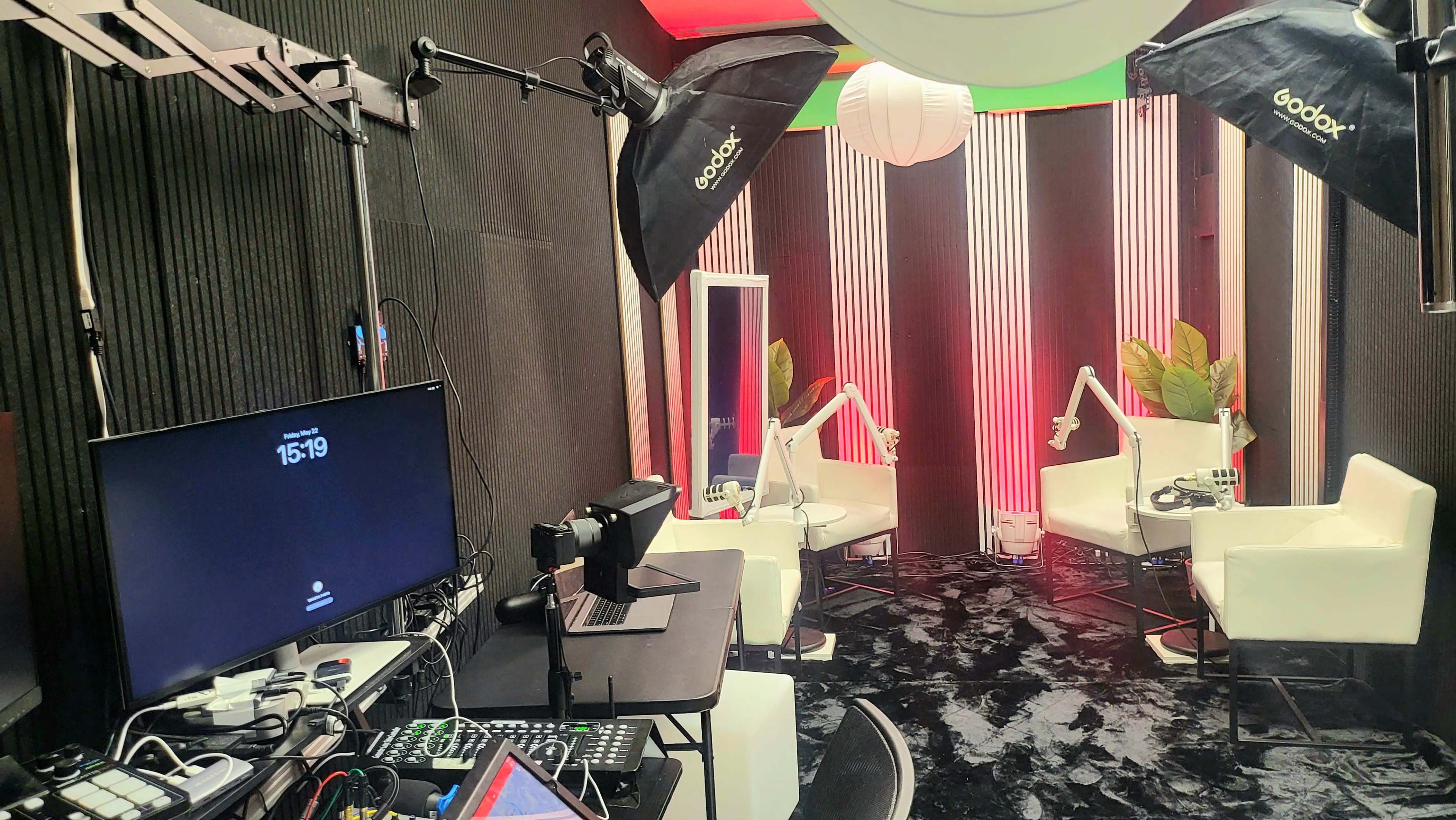 The image shows a modern studio setup with two white chairs, a large monitor displaying the time, and several lighting equipment fixtures against a backdrop of dark walls and colorful accents.