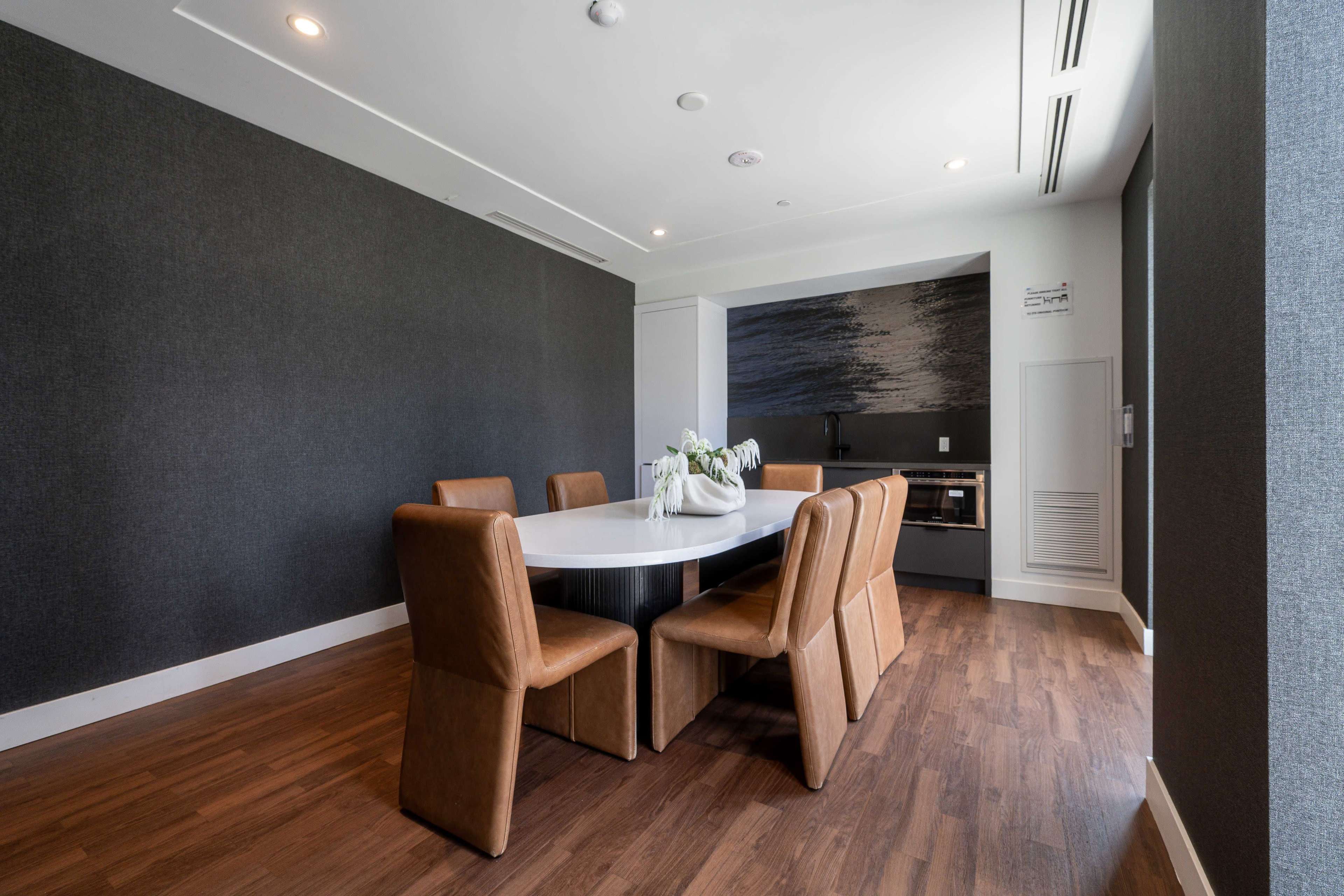 The image shows a modern dining area featuring a round table surrounded by brown leather chairs and a minimalist kitchen in the background.