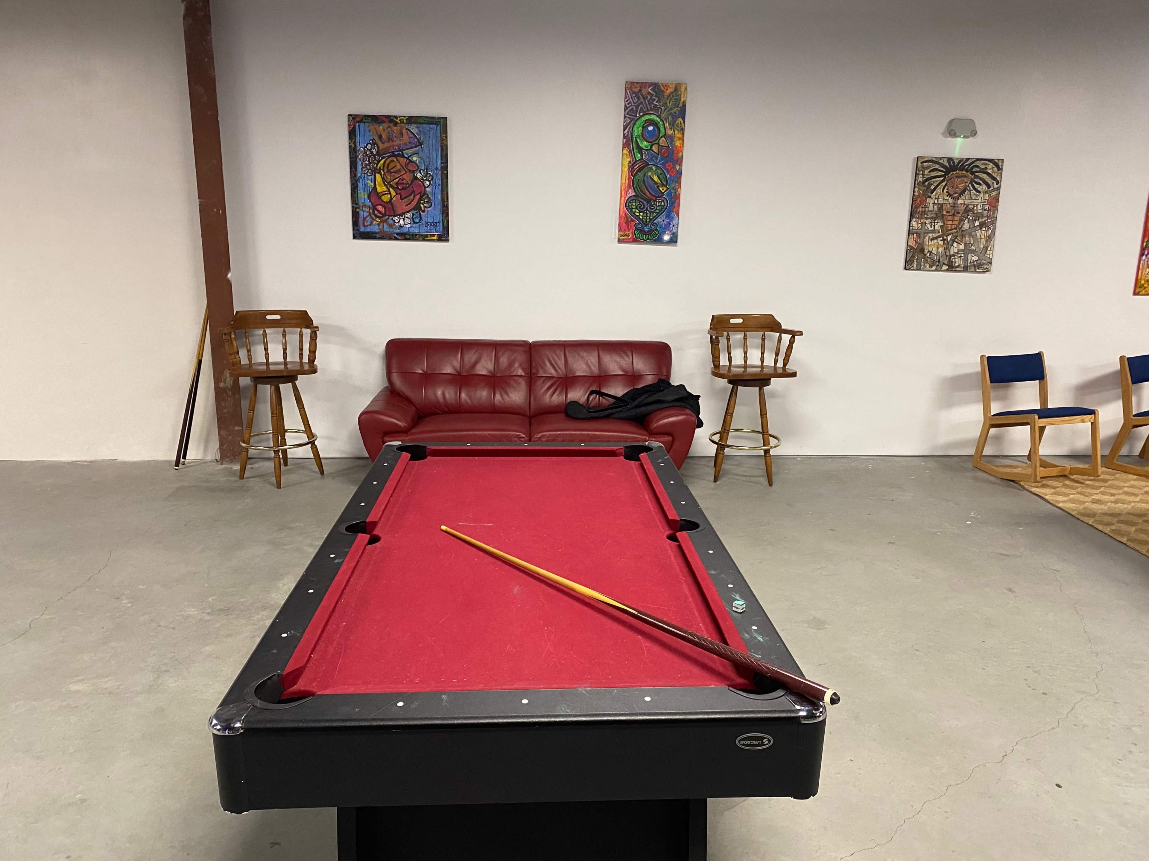 A red pool table with a cue stick rests on it in a room featuring a red sofa, bar stools, and colorful wall art.