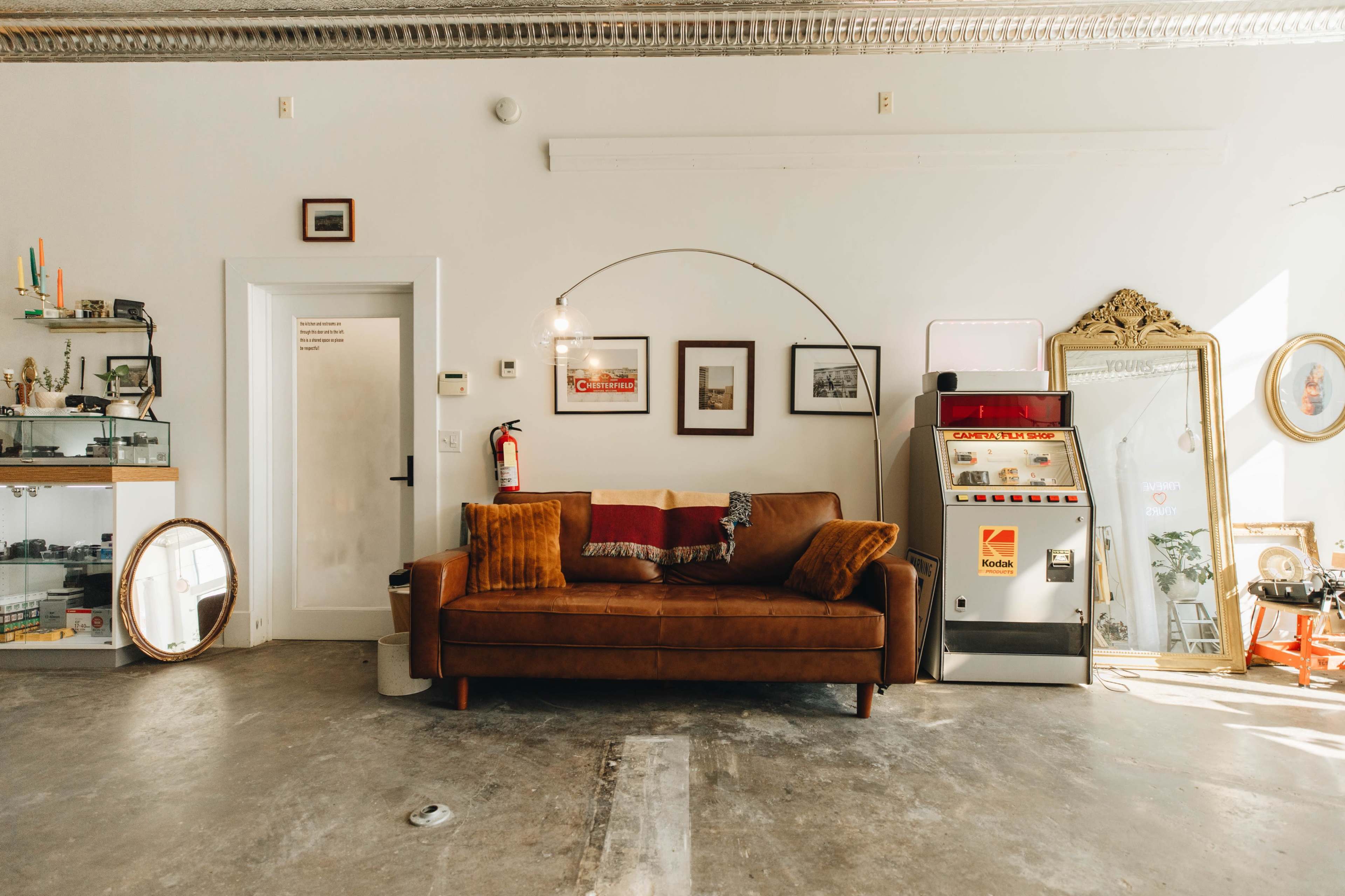 A brown leather couch with decorative pillows sits facing a wall adorned with framed pictures and an antique mirror, next to a vintage Kodak photo booth.