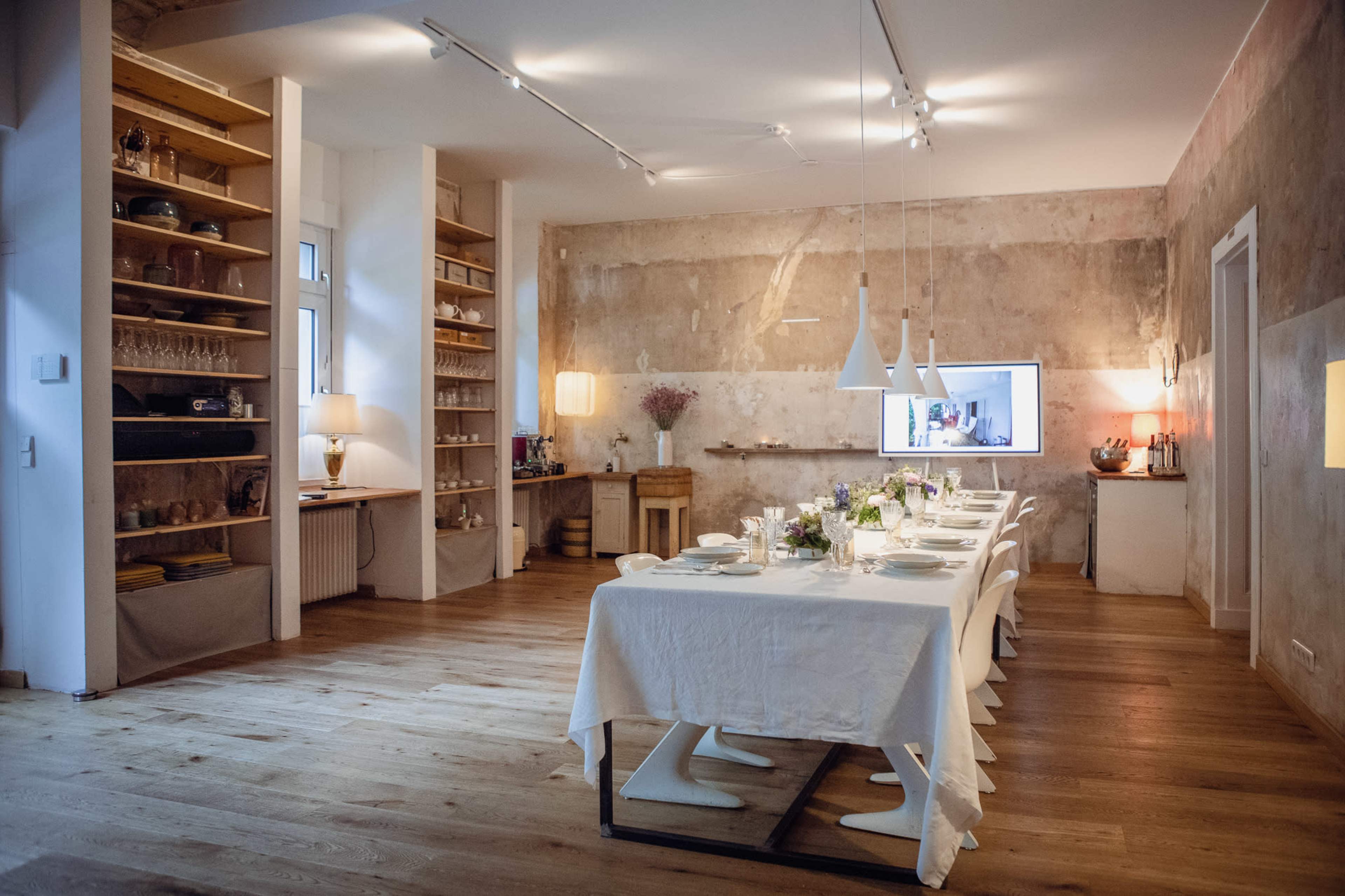 A long dining table covered with a white tablecloth is set in a room with light wood flooring, featuring exposed walls and shelves filled with dishware and glasses.