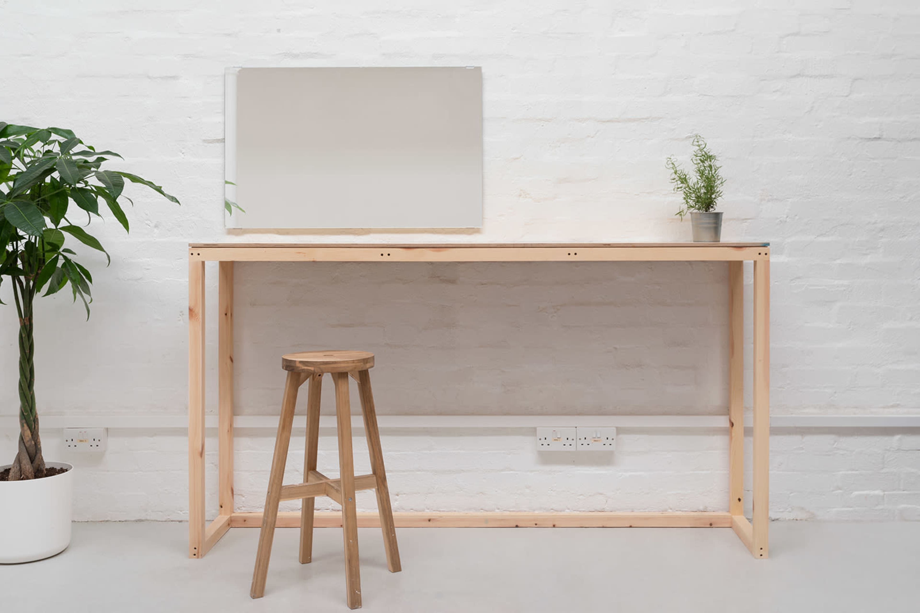 A wooden table with a mirror above it and a stool is positioned against a white-brick wall, alongside a potted plant.