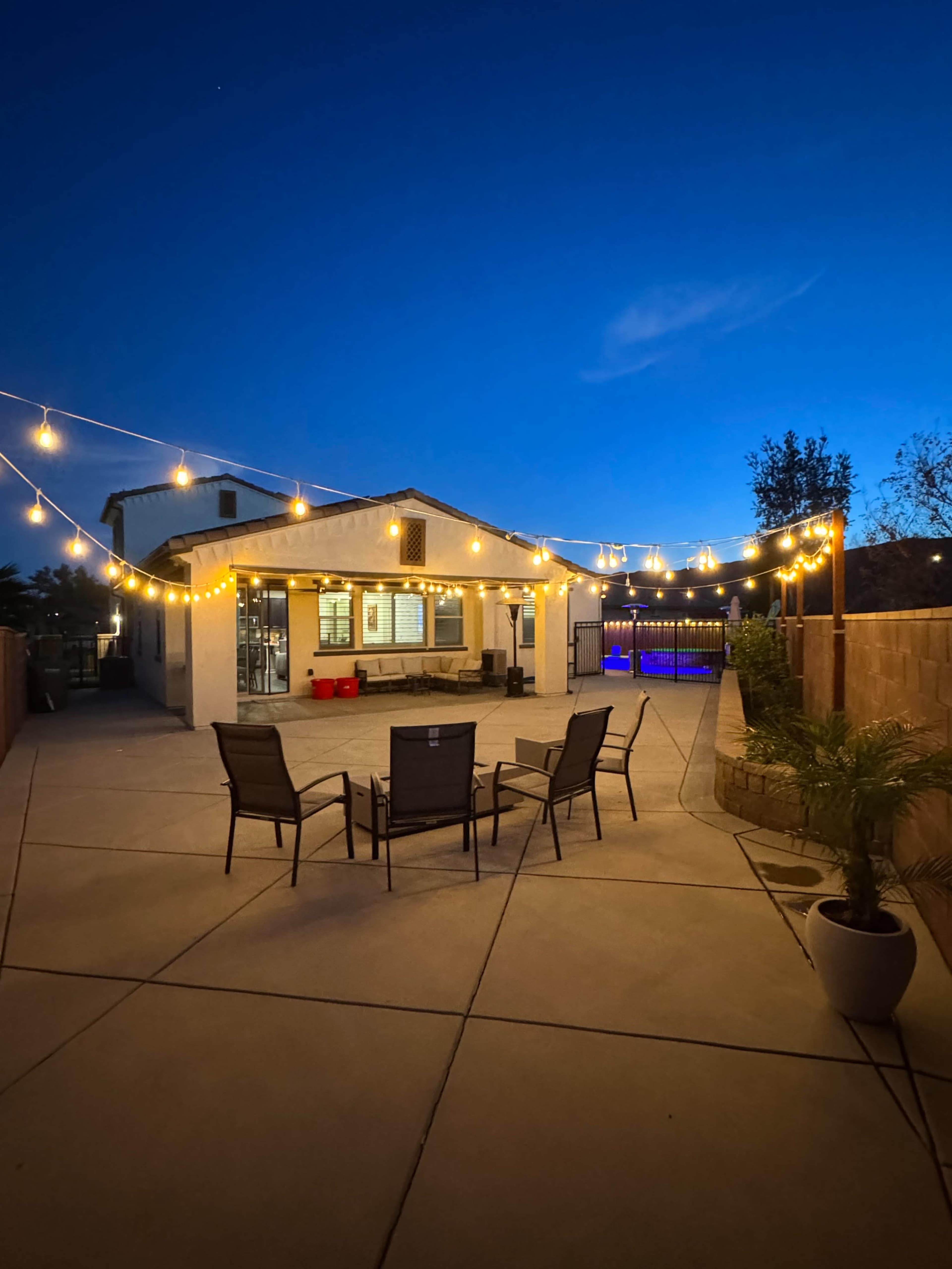 A patio area with four chairs is illuminated by string lights against a twilight sky, featuring a house and a pool in the background.