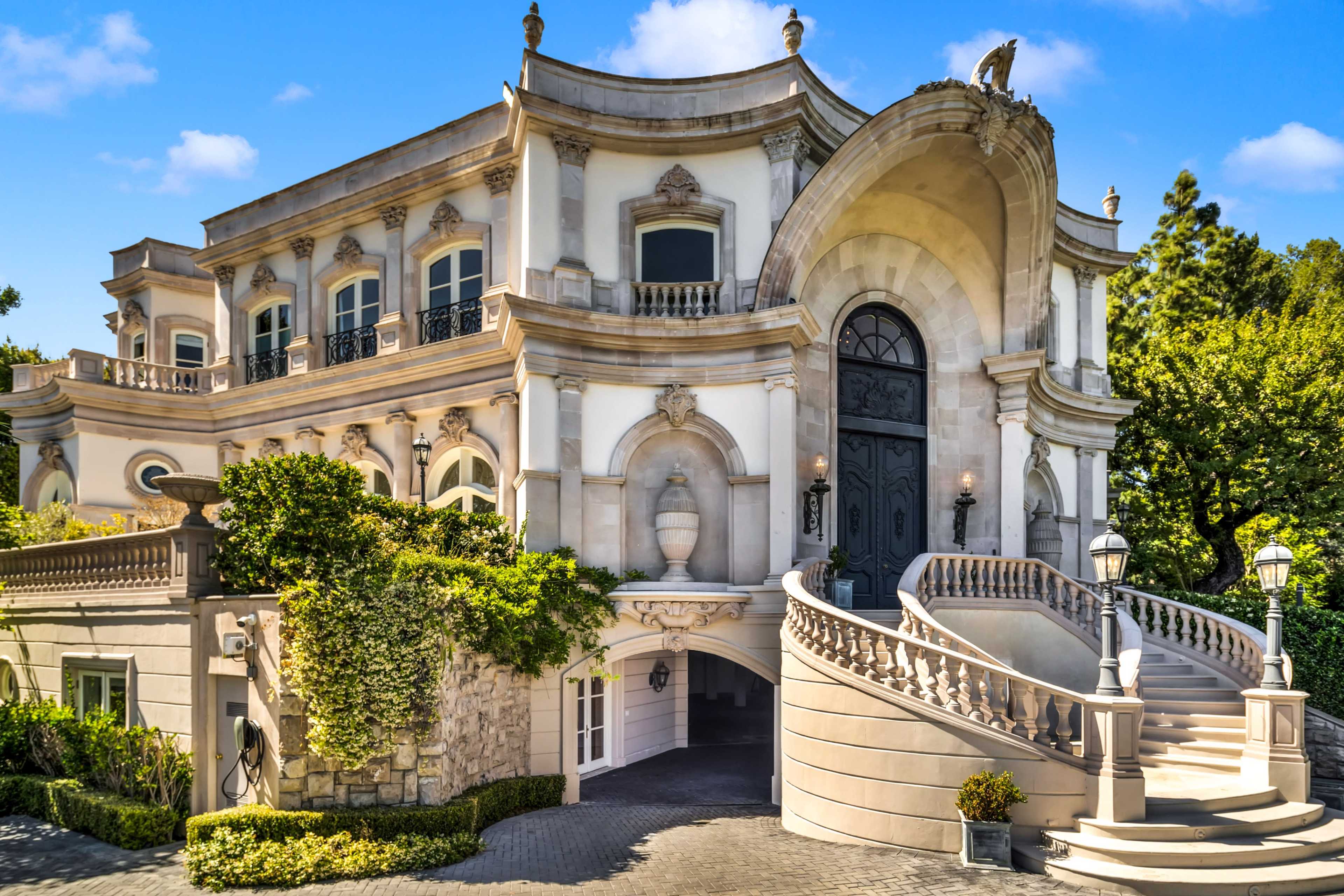 A large, ornate mansion with a grand entryway, intricate architectural details, and lush landscaping is set against a clear blue sky.