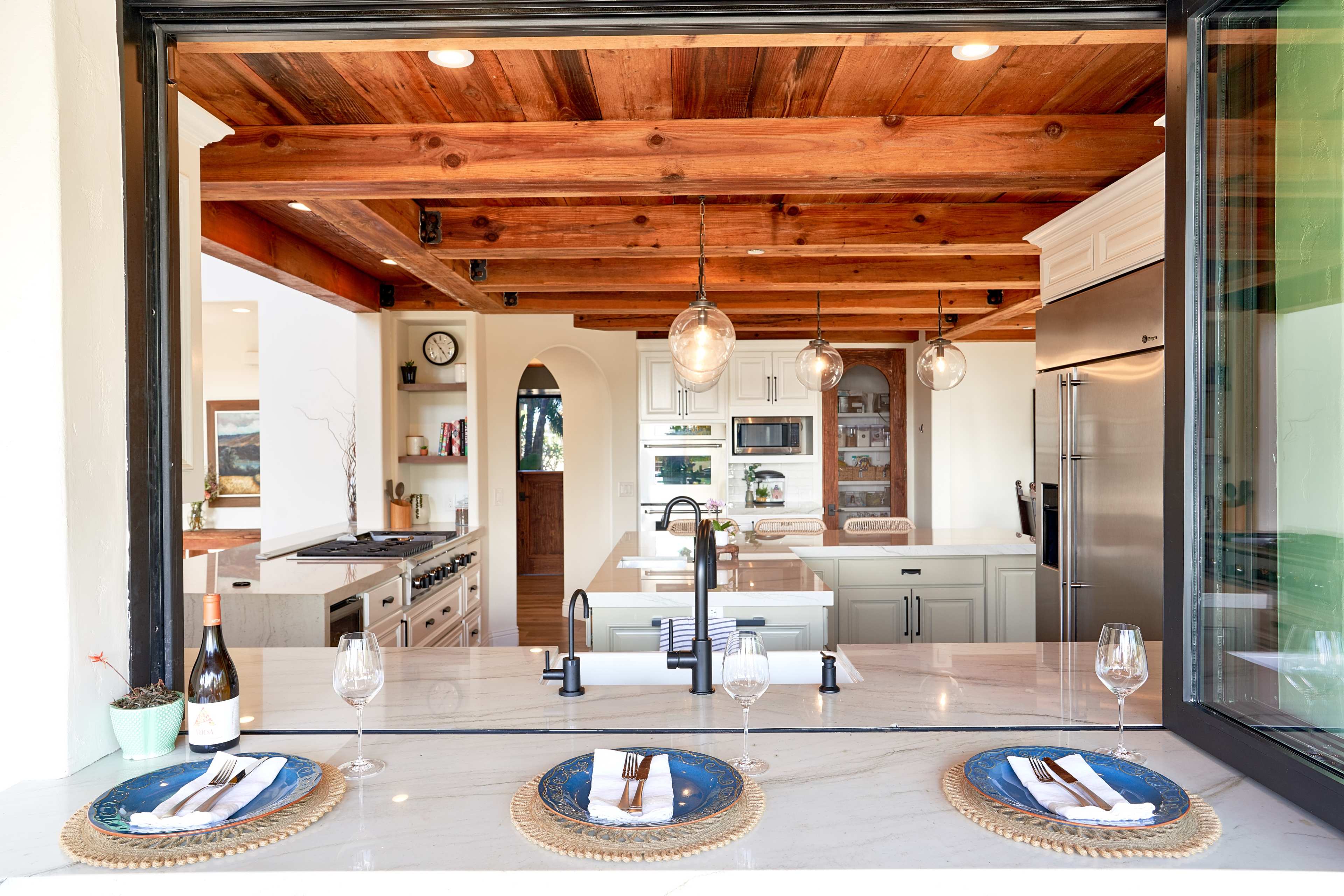 A modern kitchen with wooden beams features an open window overlooking a dining area set with plates and wine glasses.