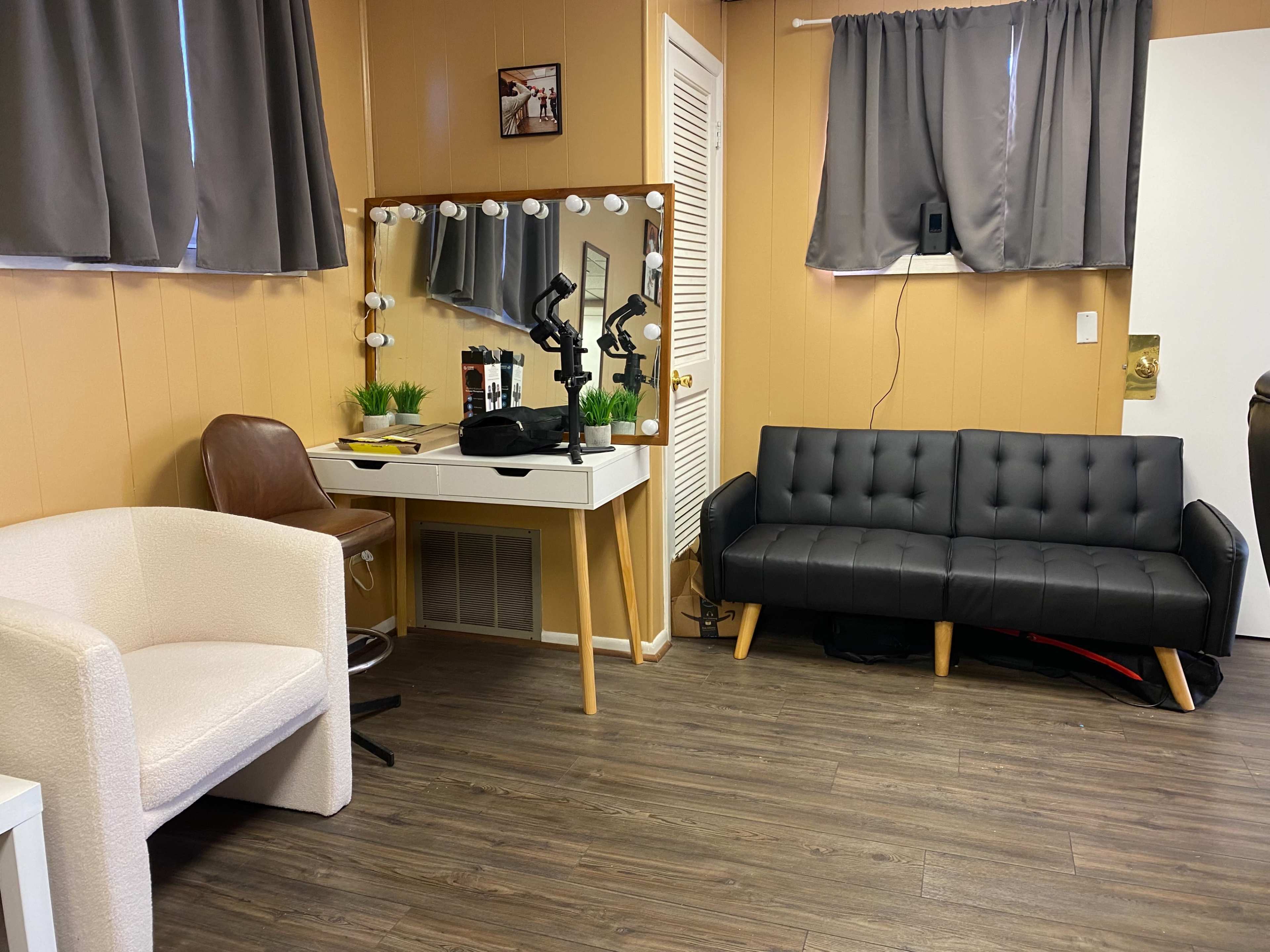 The image shows a small room with wooden flooring, featuring a black couch, a white chair, a vanity table with a mirror and lights, and decorative plants.