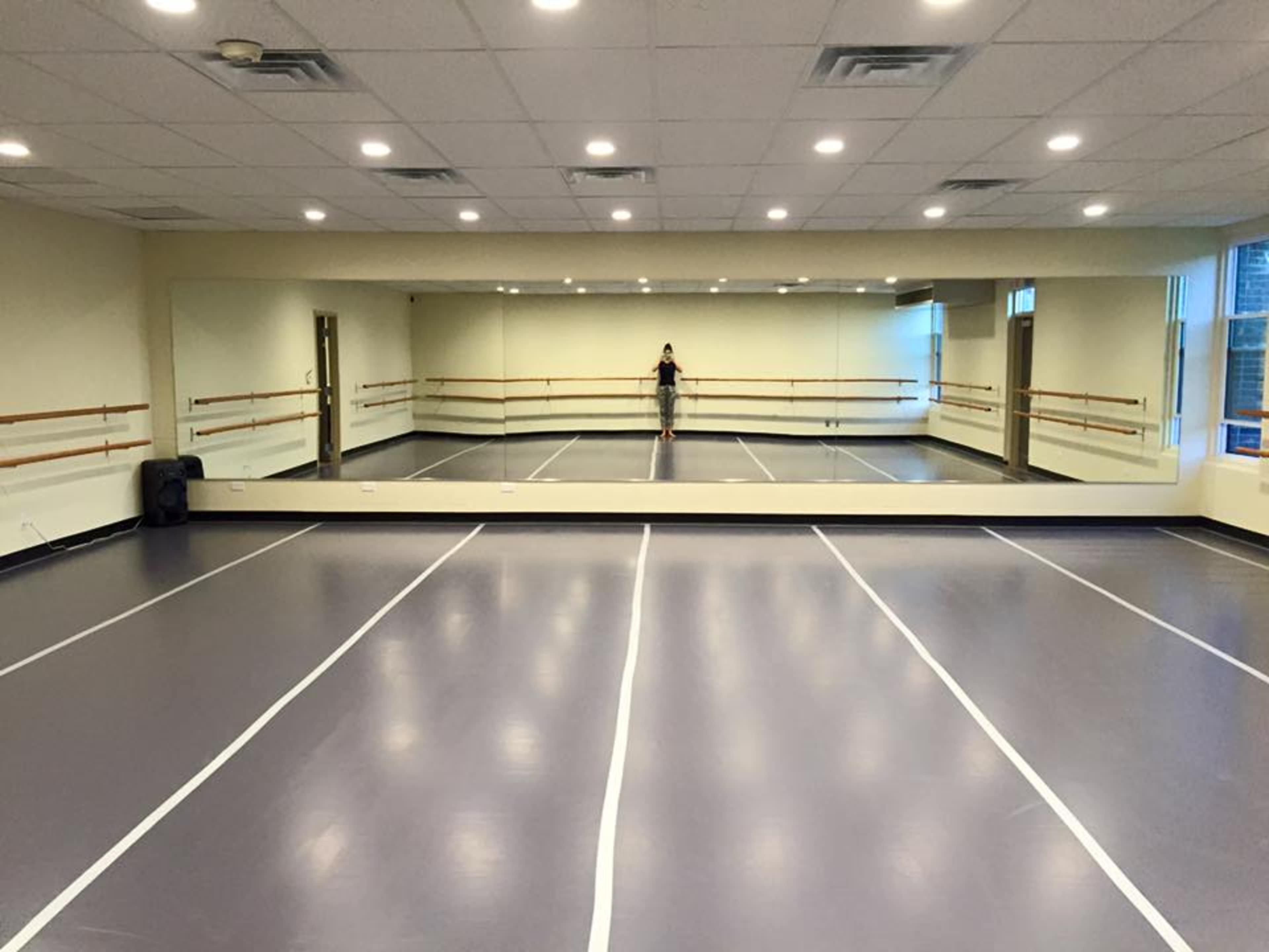 A spacious dance studio features a mirrored wall, barre along the sides, and a gray floor with white marking lines.