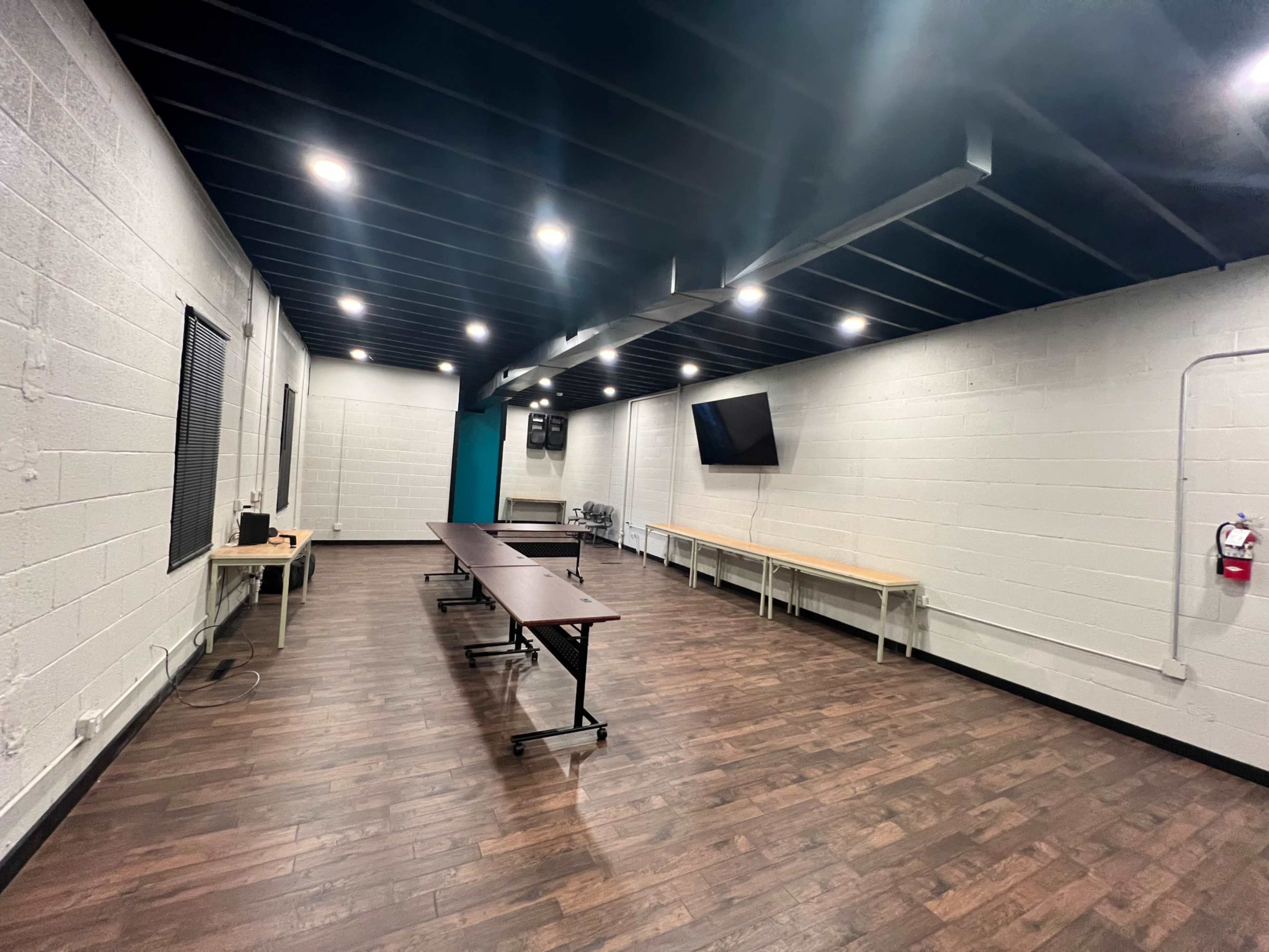 A spacious room with wooden flooring, featuring two long tables, a television mounted on the wall, and several overhead lights.