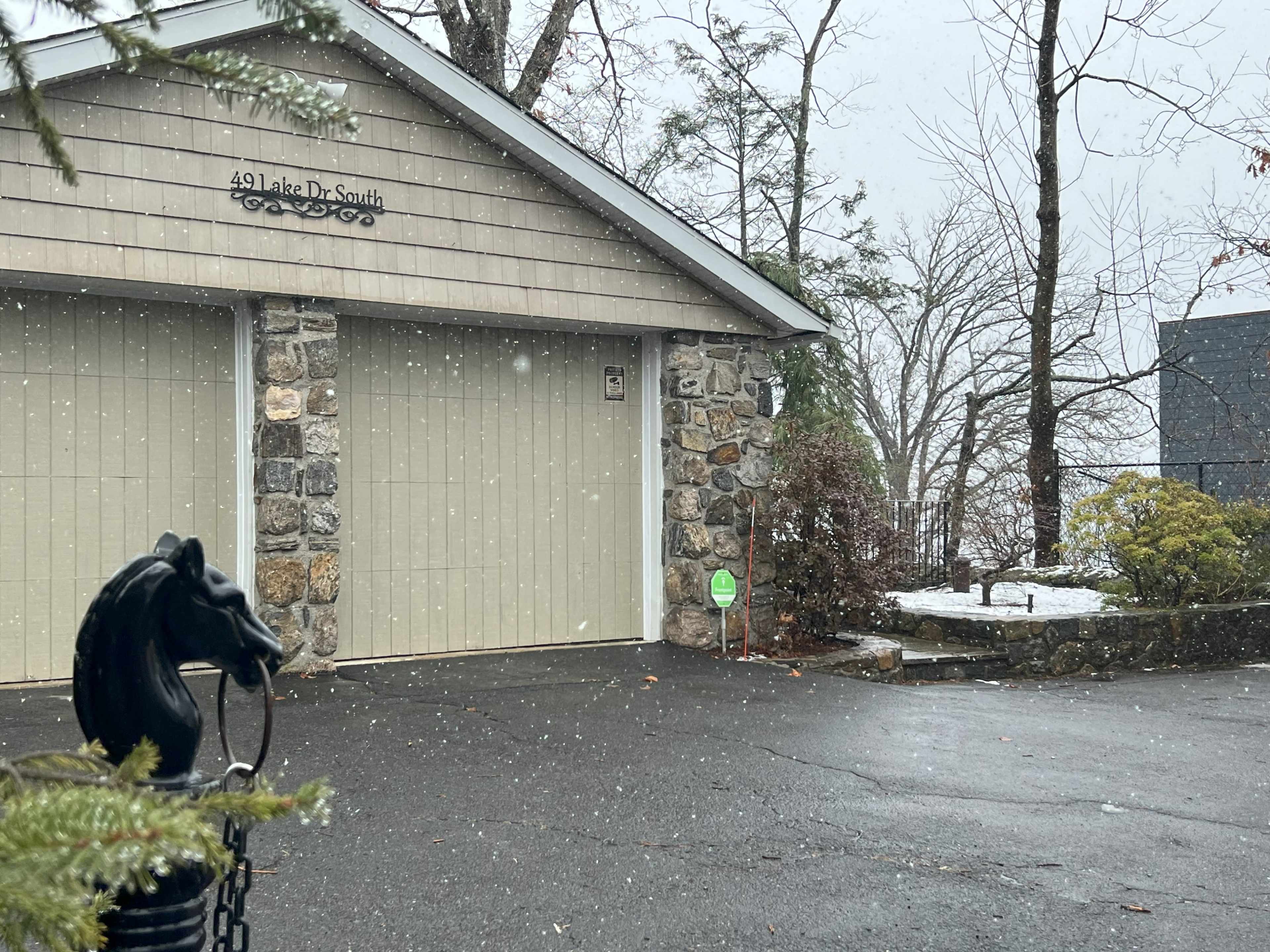 A stone and wood garage is visible, with snow falling and a horse sculpture on a black post in the foreground.