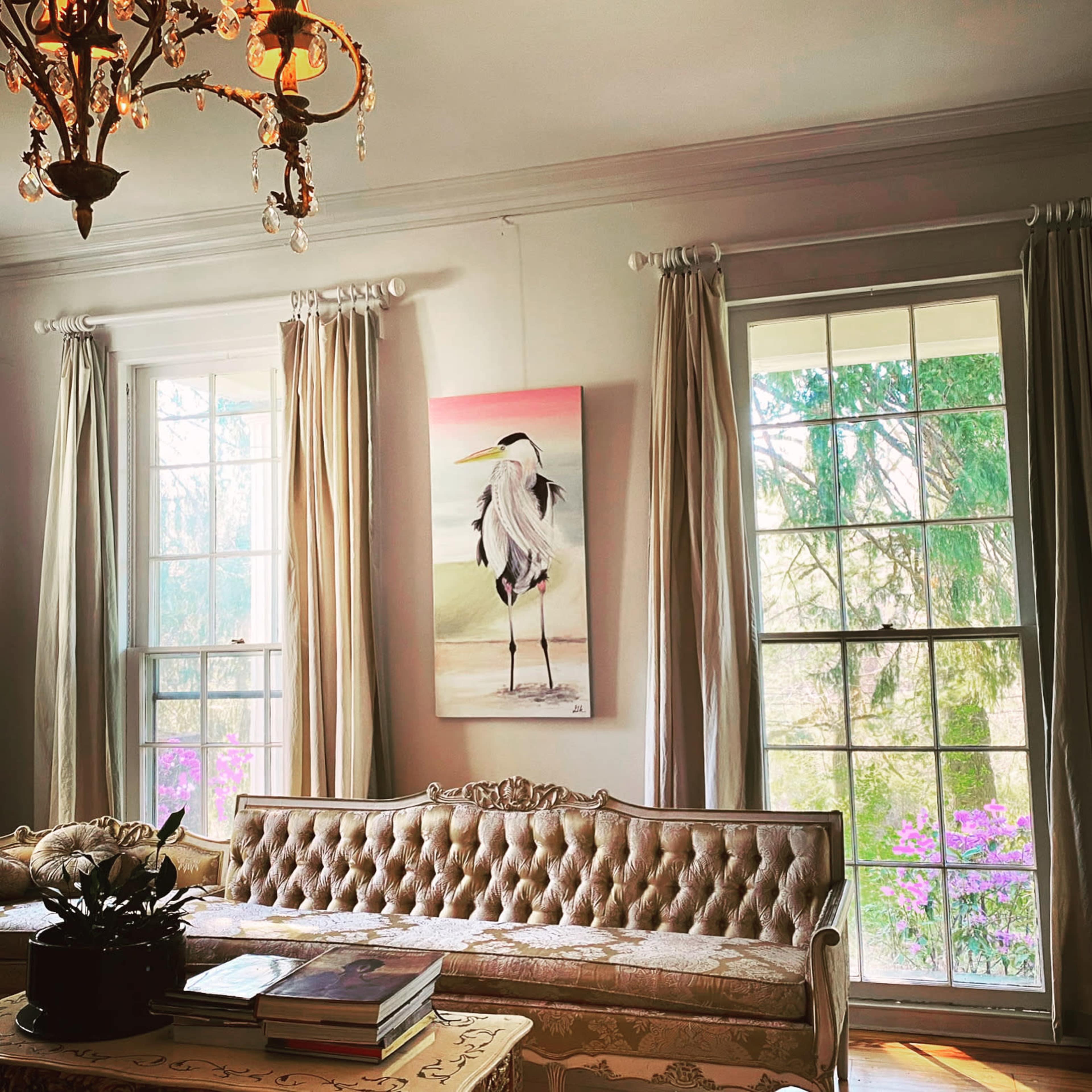 A light-filled room with a chandelier, a framed heron painting on the wall, and a sofa in front of large windows adorned with curtains.