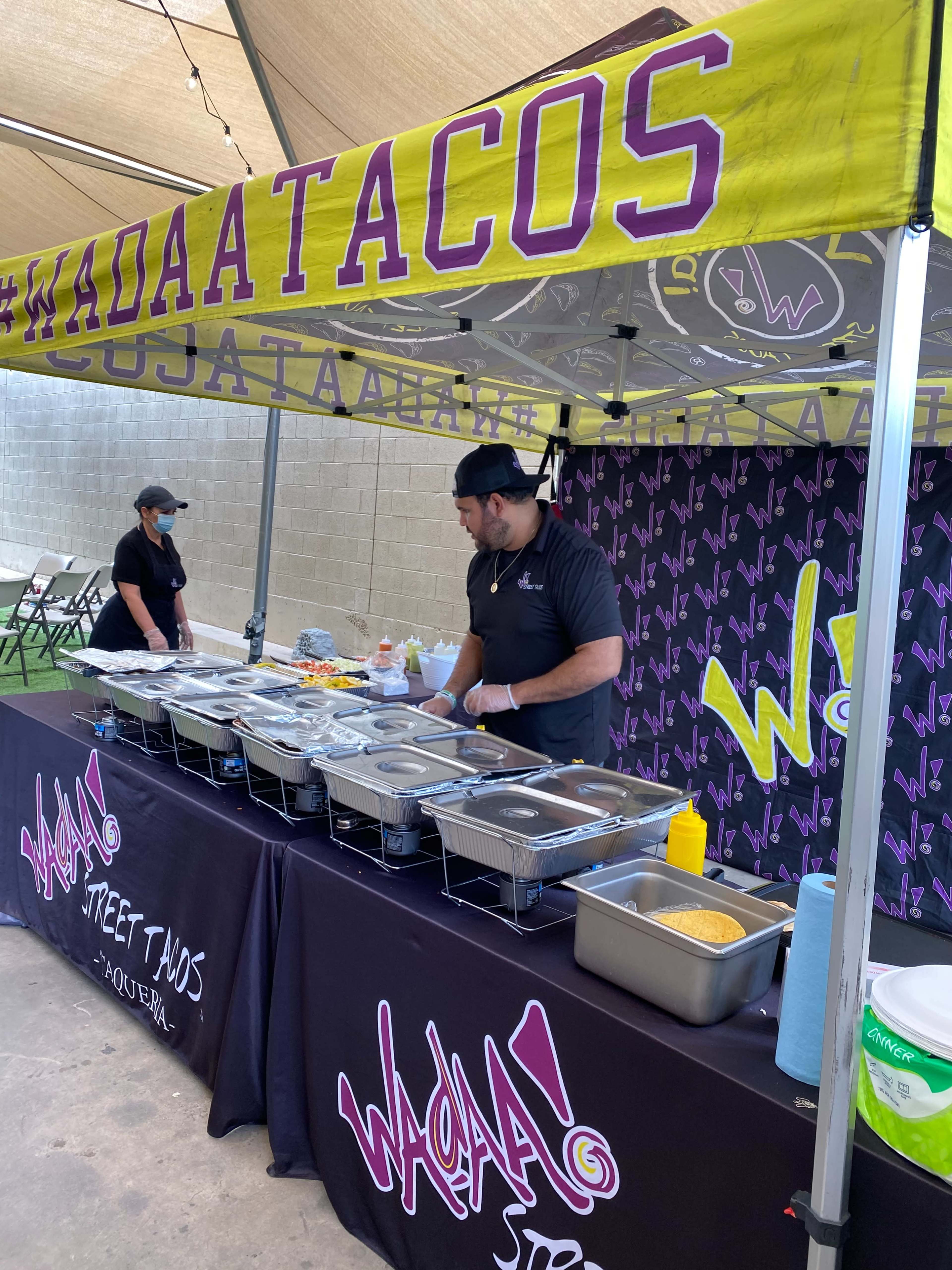 A vendor prepares food at a taco stand featuring a colorful canopy and multiple serving trays.