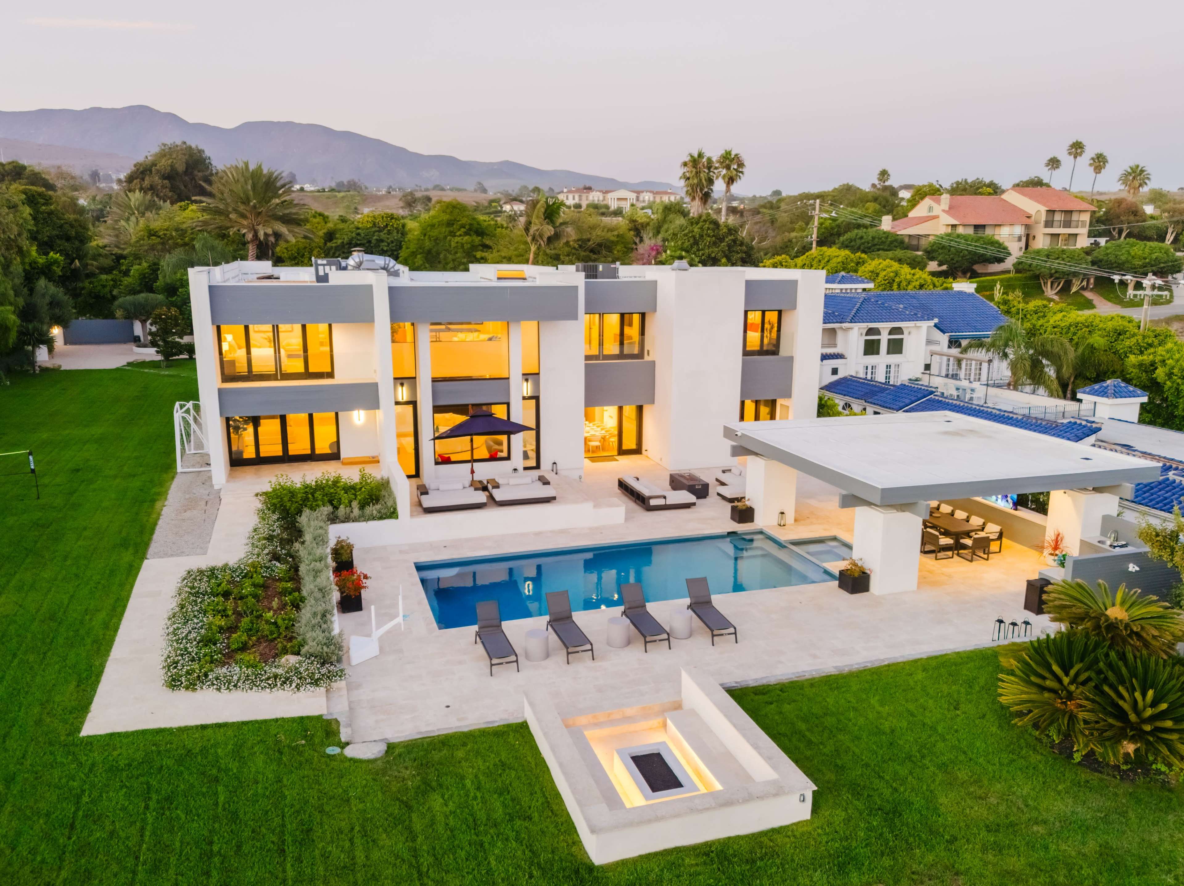 A modern two-story house with large windows, a swimming pool, and a landscaped yard is located in a suburban area with mountains in the background.