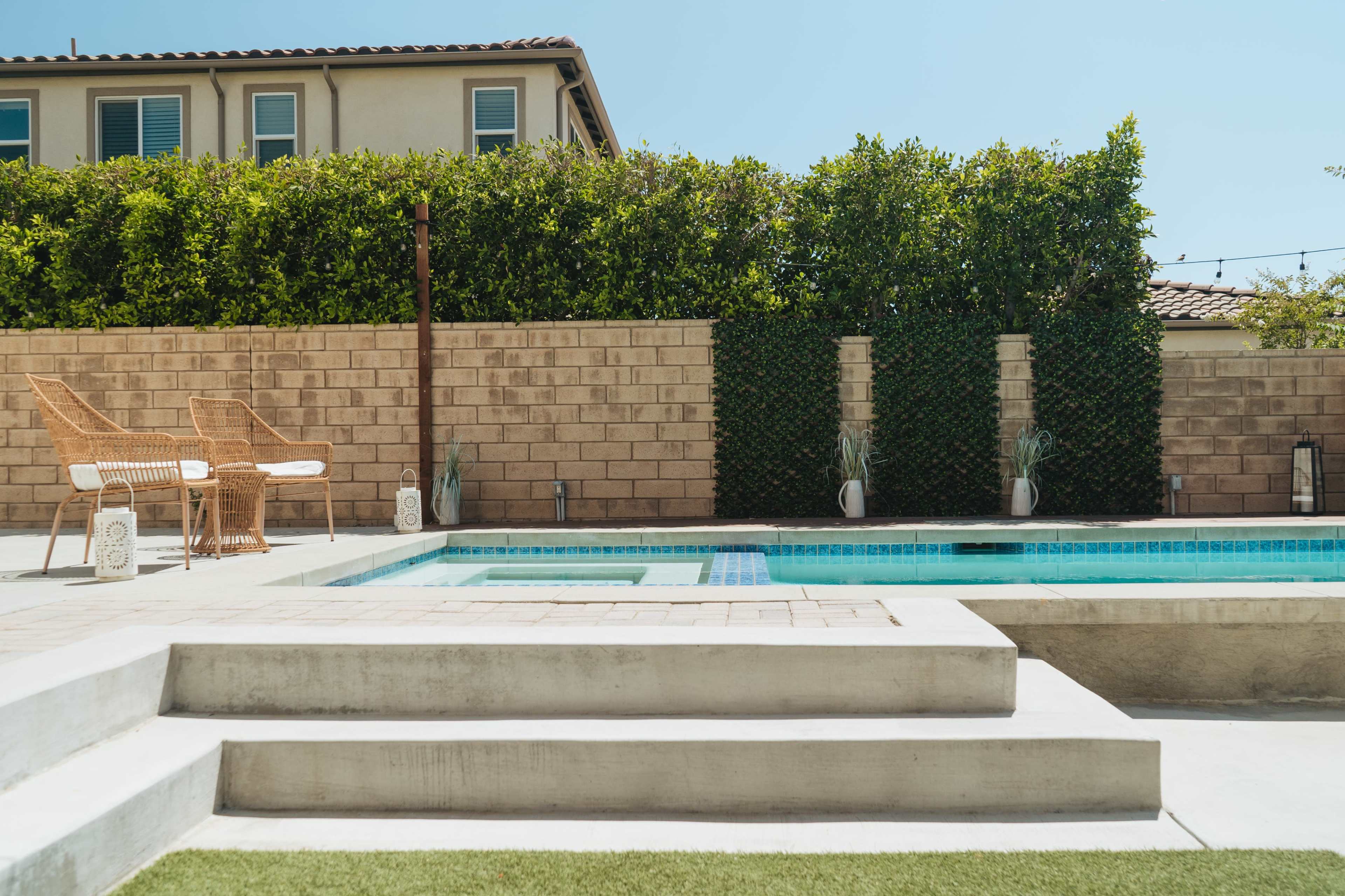 A modern backyard with a swimming pool, surrounded by a stone wall and green hedges, featuring two wicker chairs on a patio and planters near the pool.